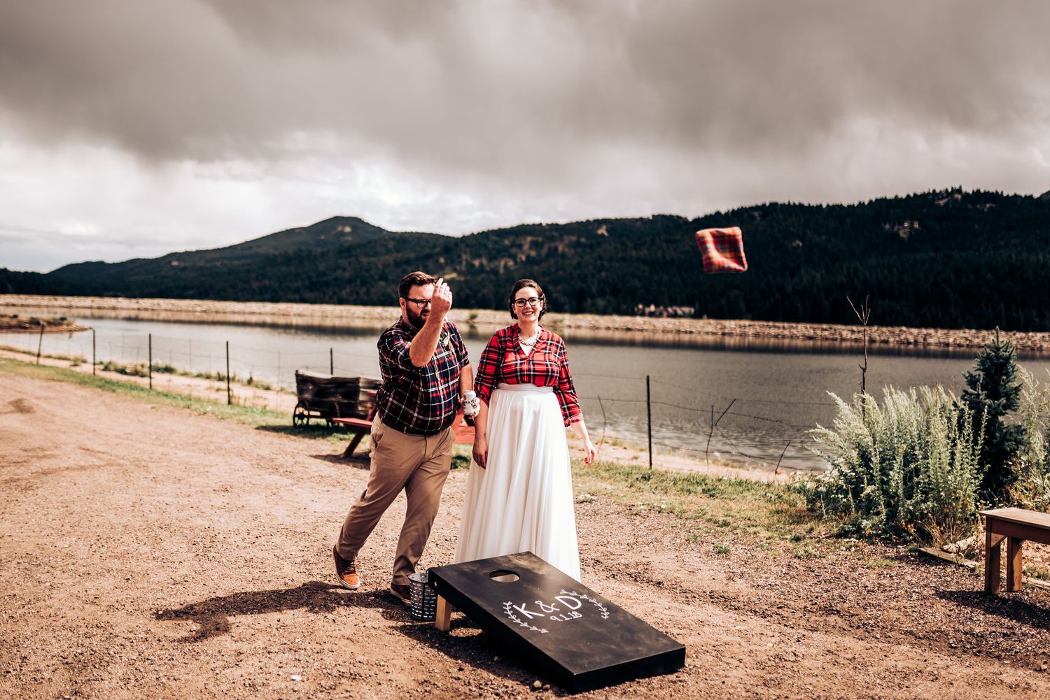 A couple playing cornhole outdoors near a lake with mountains in the background.