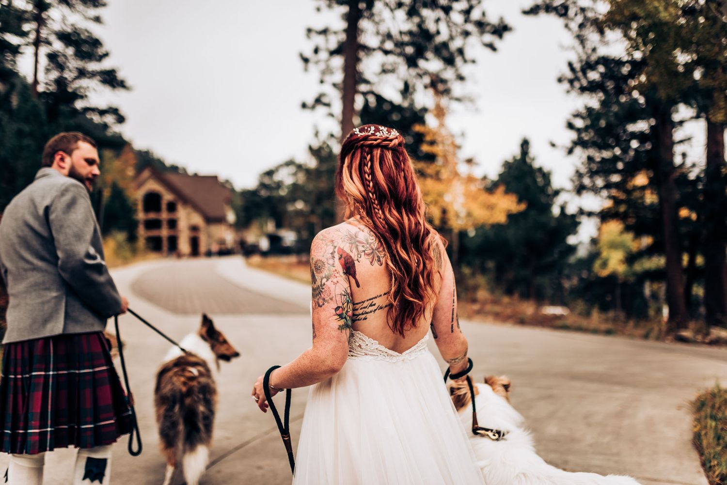 A couple in wedding attire walks their dogs along a mountain path near a rustic building.