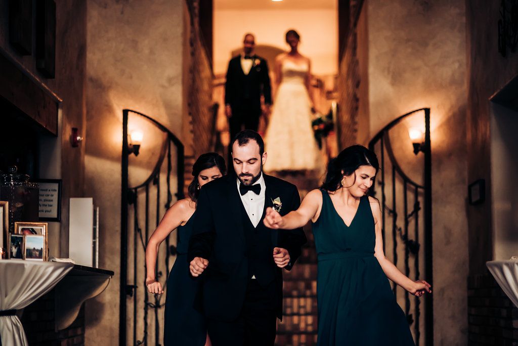 Wedding party descends elegant staircase in rustic venue with warm lighting and brick walls.