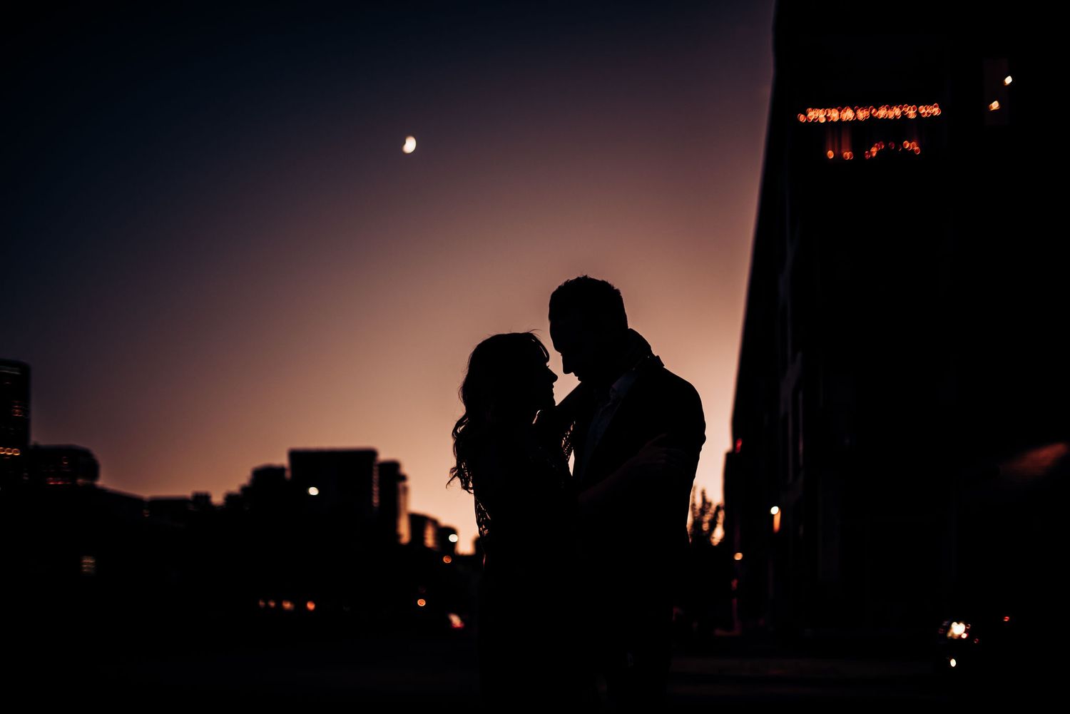 Silhouetted figures embrace against a dusky cityscape with a crescent moon overhead.