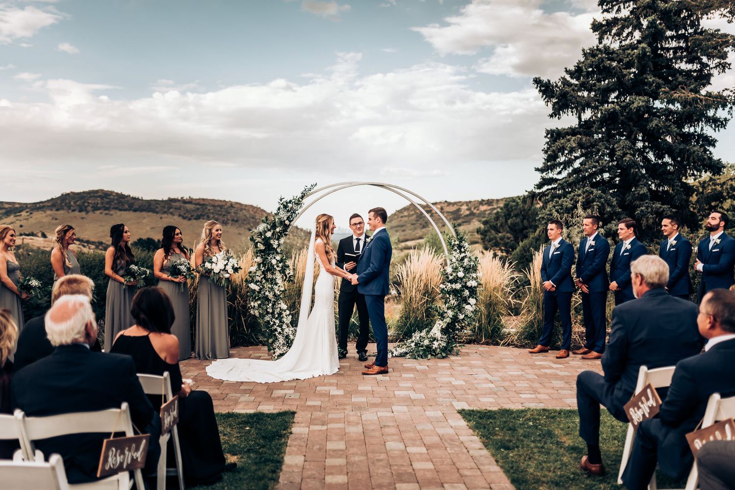 Newlyweds celebrate under floral arch as wedding guests applaud at scenic mountain venue.