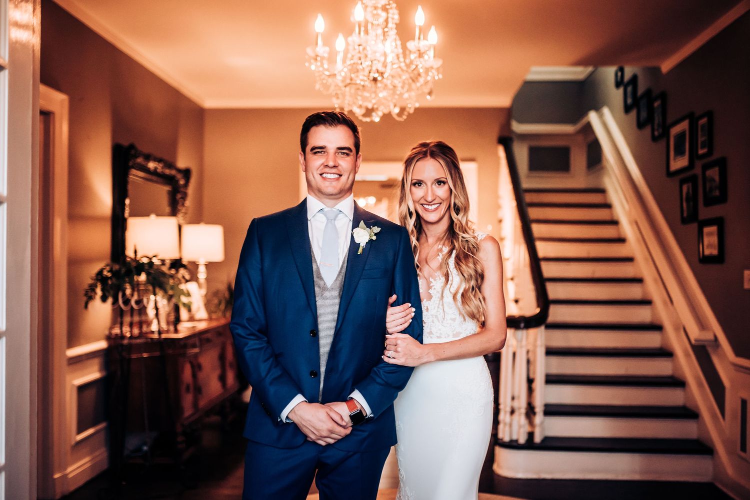 A wedding couple poses in an elegant home interior with chandelier and wooden staircase.