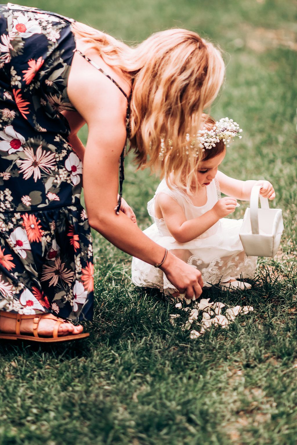 Woman in floral dress helps young flower girl with basket on grassy lawn.