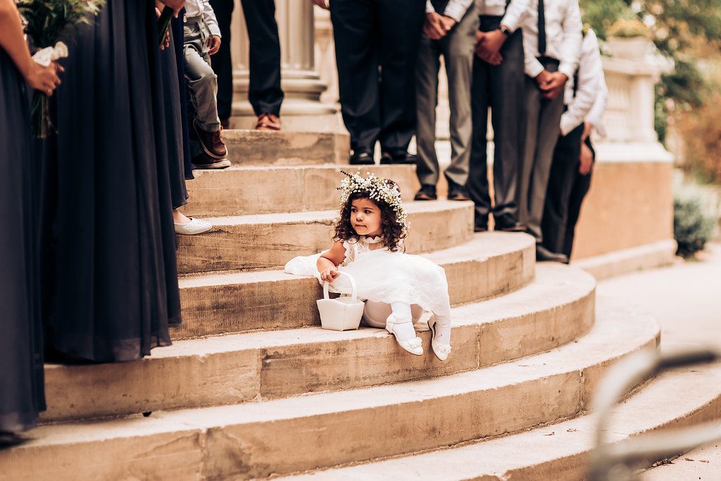 A small attendant in white sits on stone steps during an outdoor wedding ceremony.