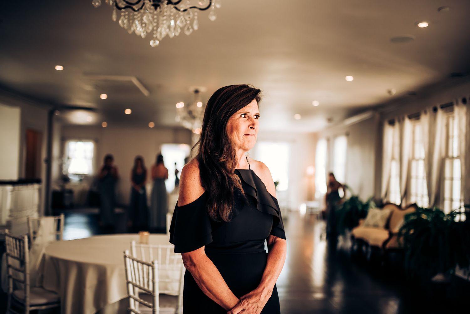 Woman in elegant black dress stands in reception venue with chandelier overhead.