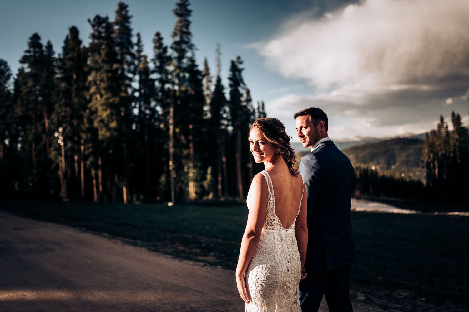 Stylish wedding couple poses together on a mountain path with evergreen trees in background.