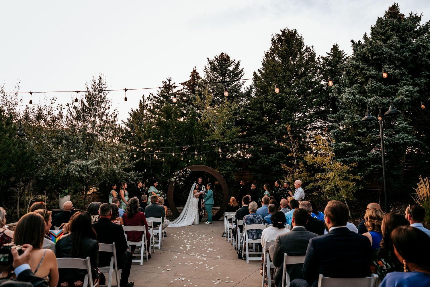 Outdoor wedding ceremony with guests seated in white chairs facing couple at floral arch amid tall trees.