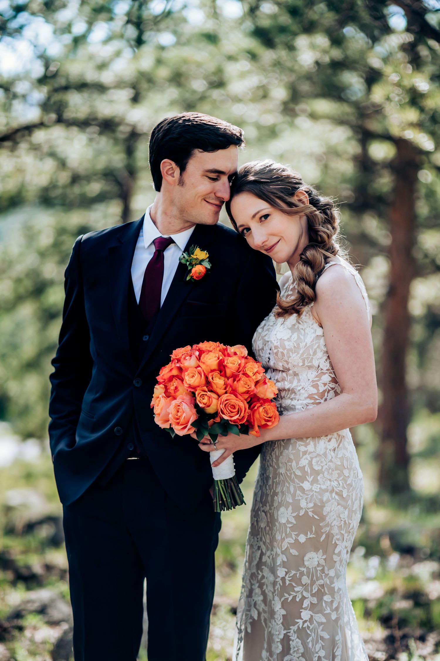 A romantic wedding couple poses outdoors with vibrant coral roses and a lace dress against a natural forest backdrop.