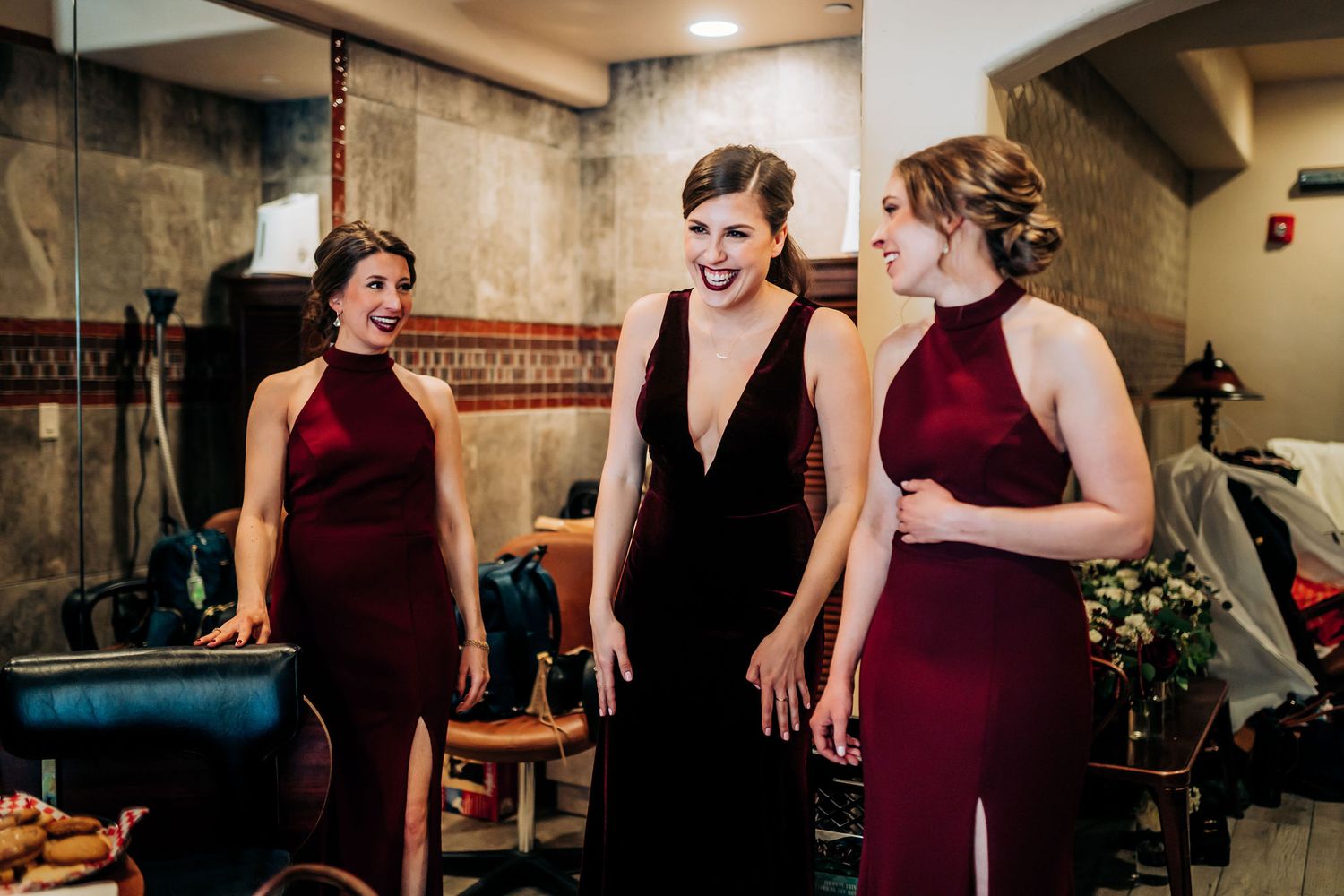 Three women in elegant dresses, two in burgundy and one in black, share a candid moment at an indoor event.