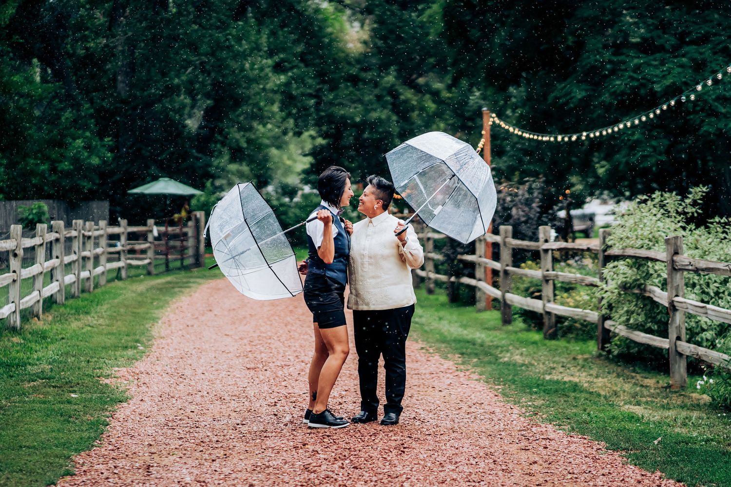 Couple posing with clear umbrellas on a rustic path with string lights.