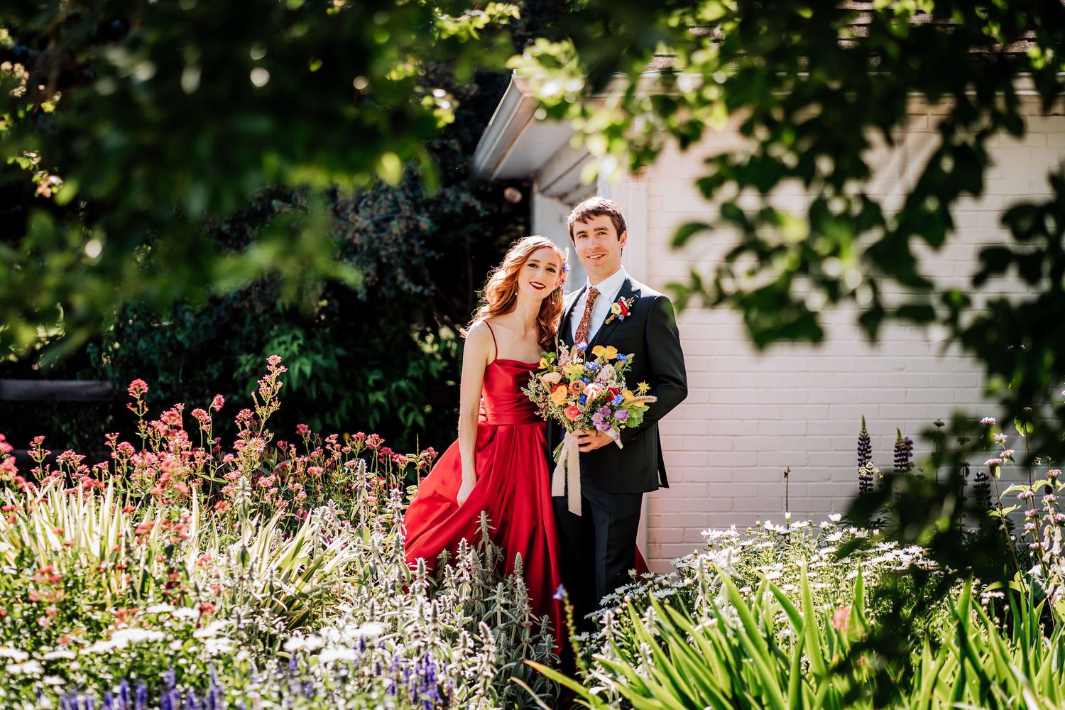 Couple in formal attire poses in blooming garden with vibrant red dress and black suit.