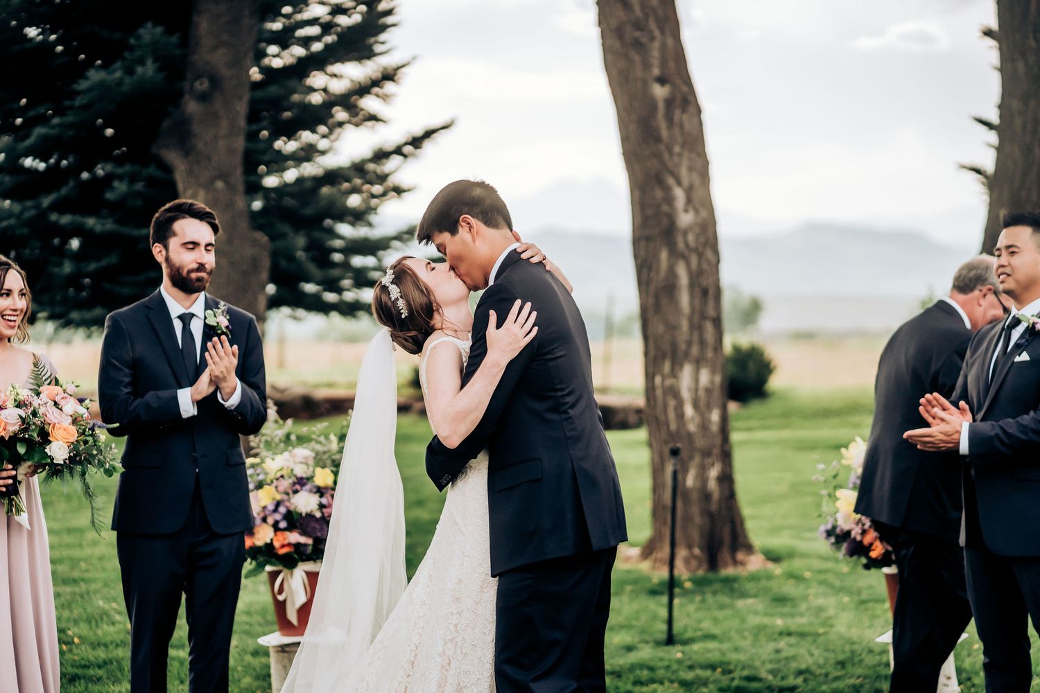 A romantic wedding kiss during an outdoor ceremony while guests and wedding party look on.