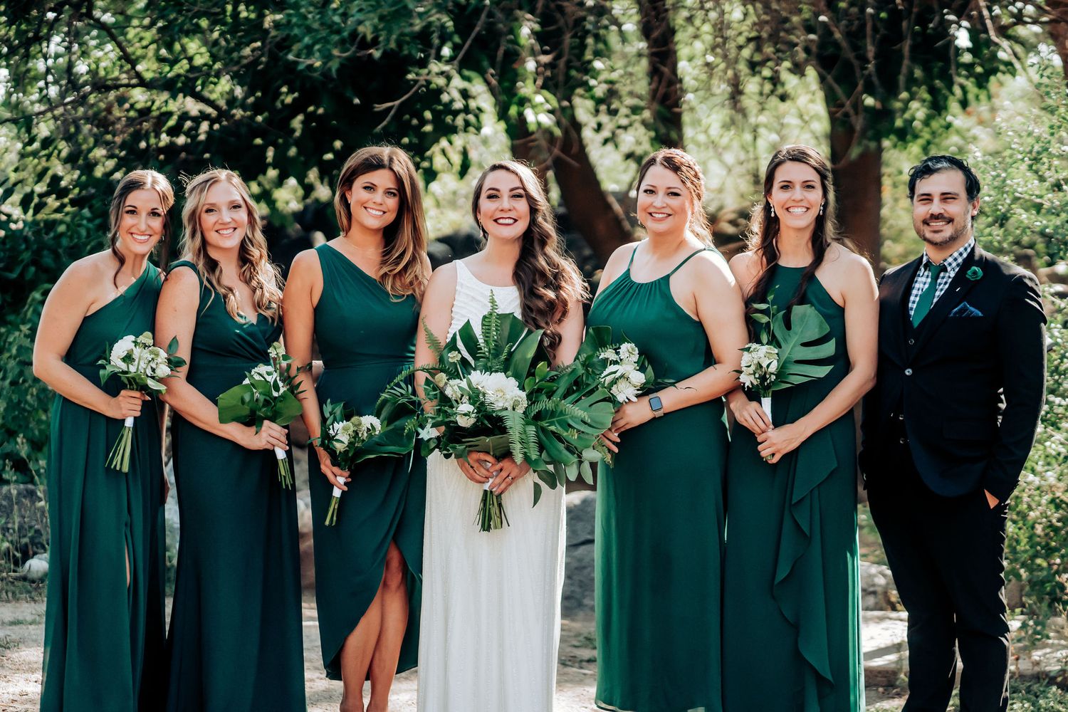 Wedding party in coordinated emerald green dresses and suits pose together in a garden setting.