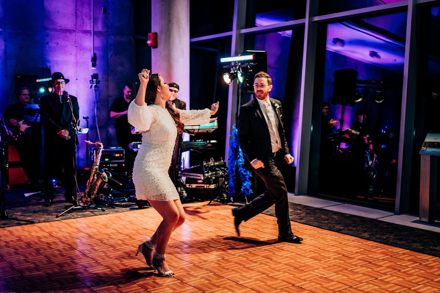 Two people dance energetically on a wooden floor during a reception with band in background.