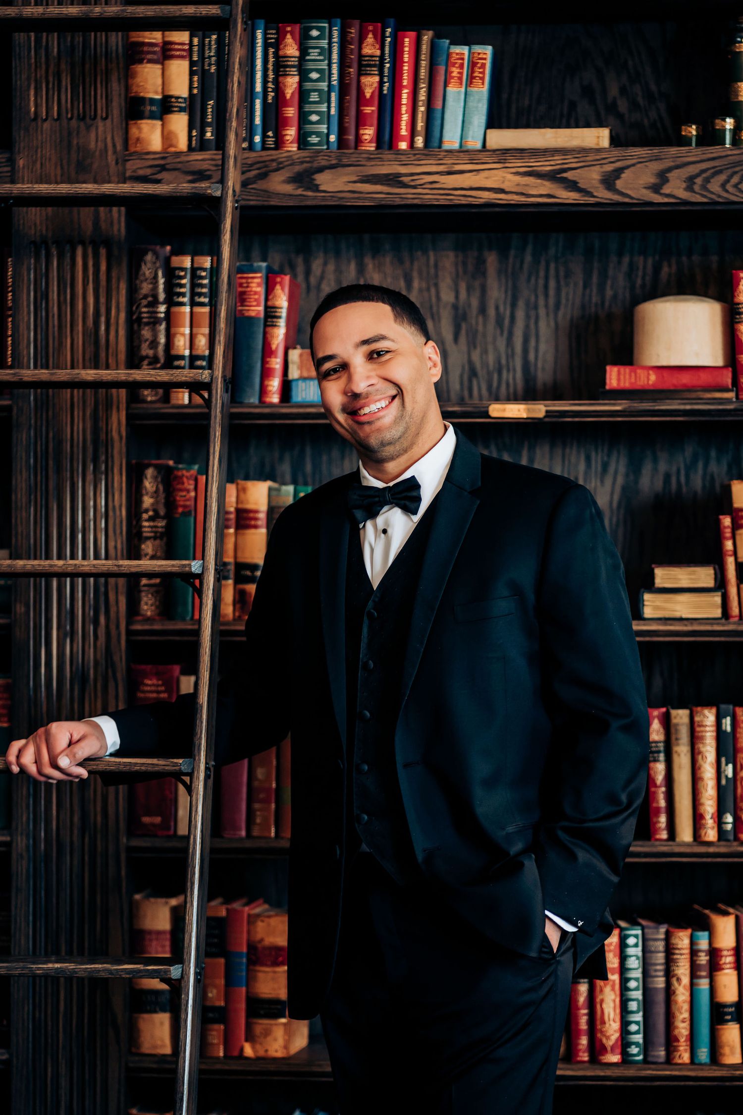 A groom in a black tuxedo poses against vintage library bookshelves.