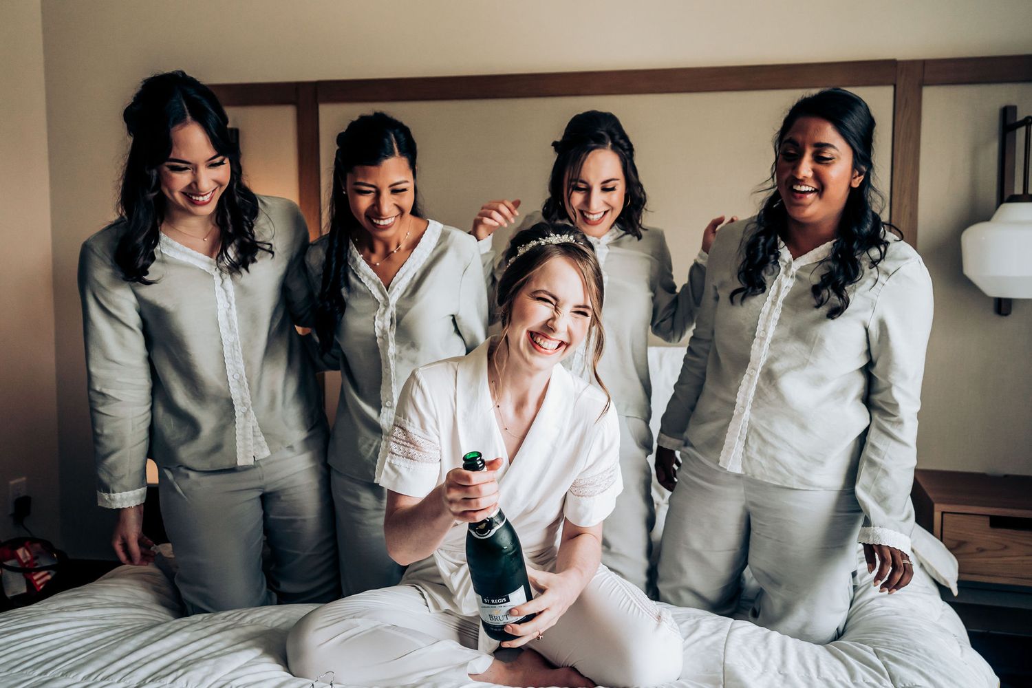 Bridesmaids in matching grey loungewear celebrate on hotel bed during wedding morning.
