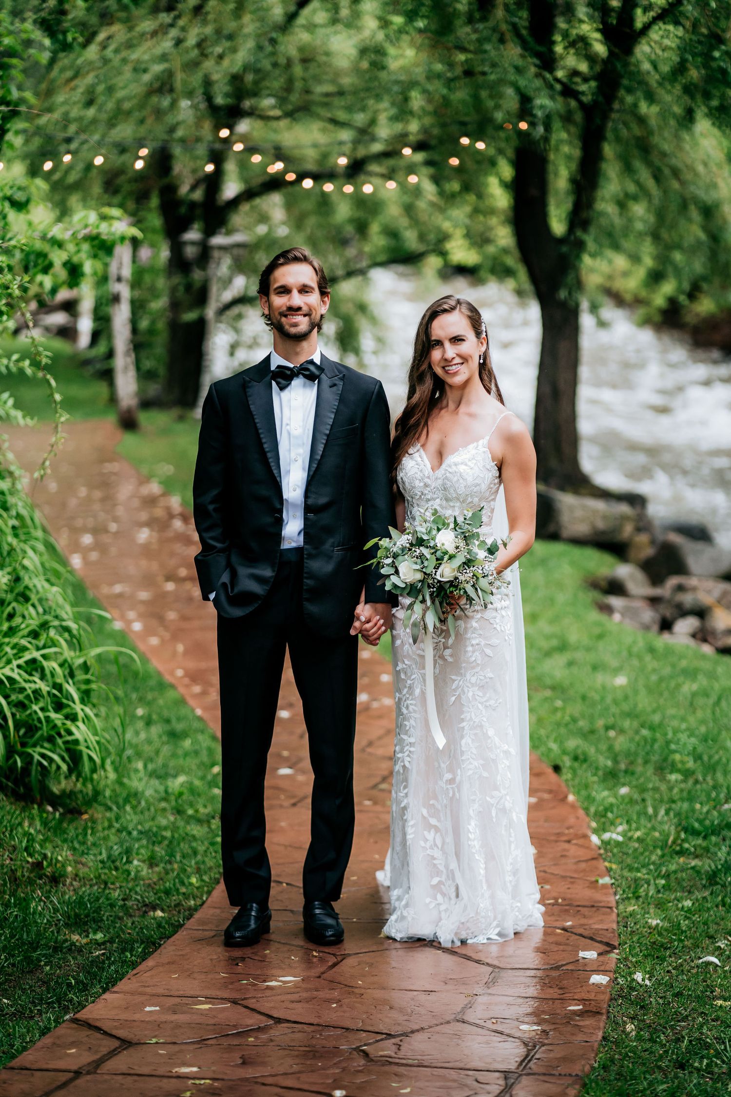 A couple poses on a stone path in their formal wedding attire with string lights and trees in the background.