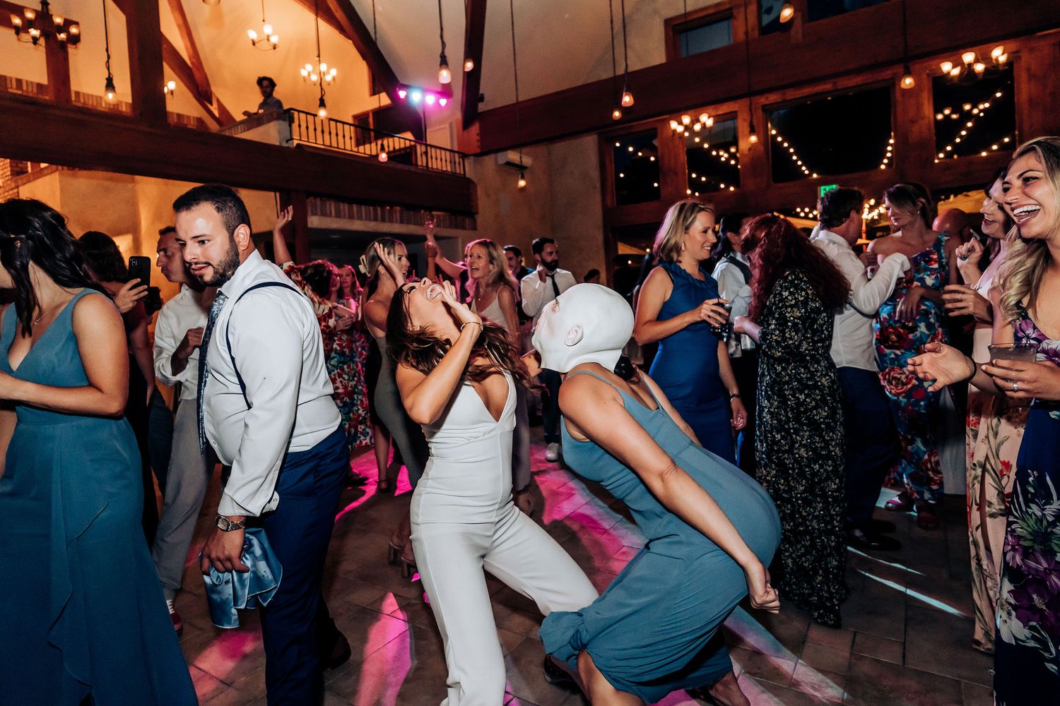 Wedding reception guests dancing in formal attire at a venue with high ceilings and mood lighting.