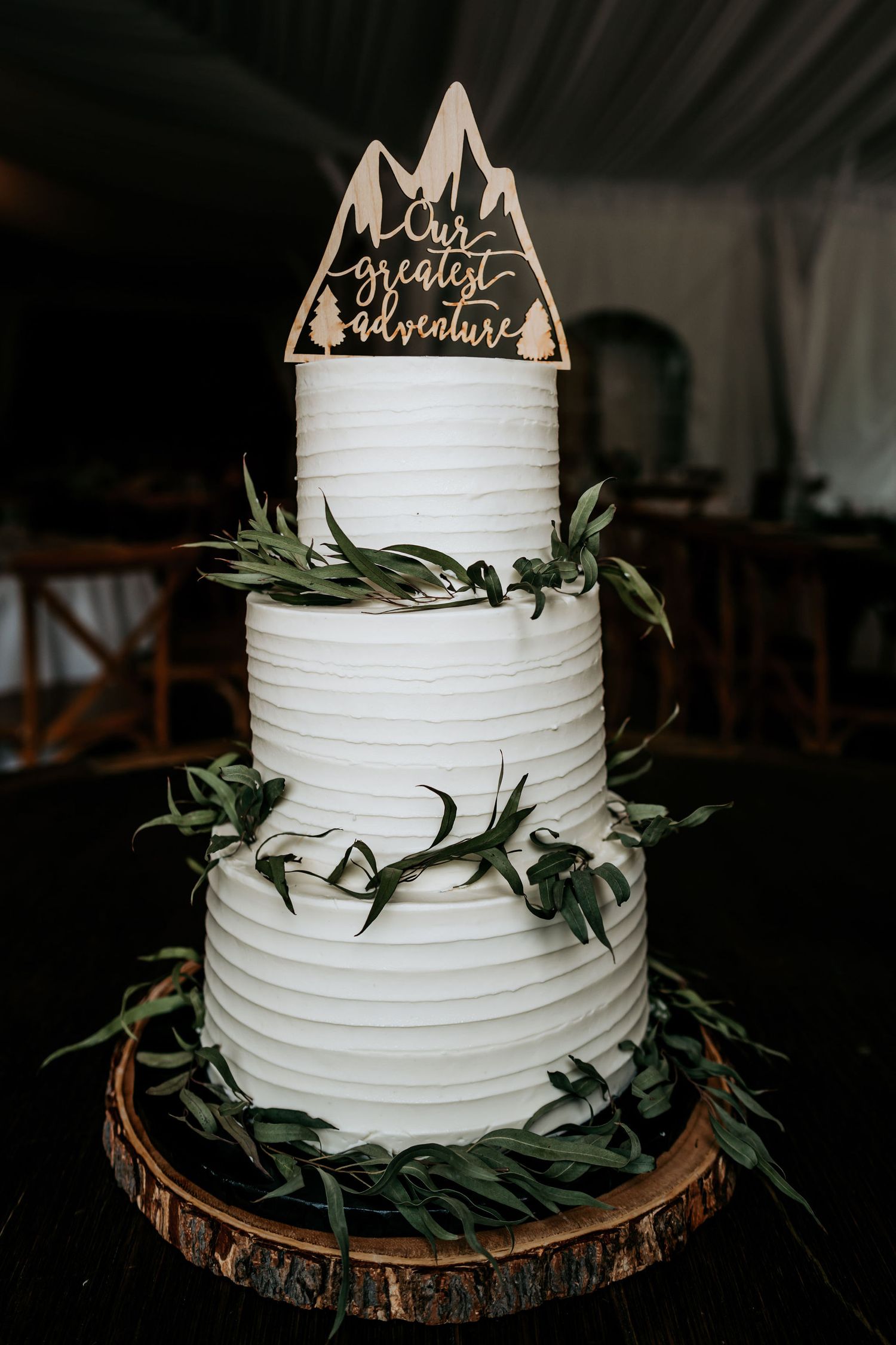 A three-tiered white wedding cake decorated with greenery and a mountain-themed topper on a wood slice base.