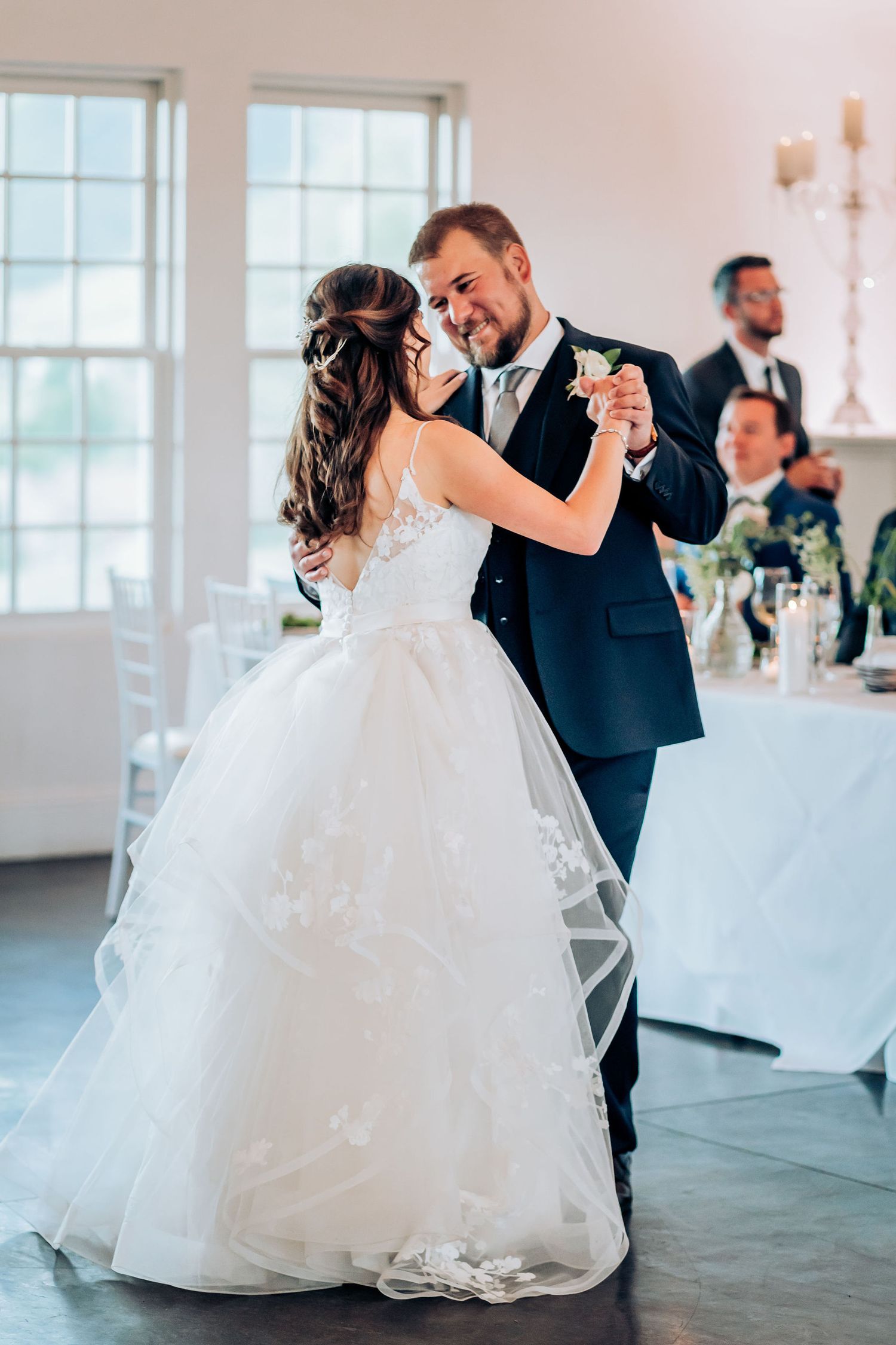 Newlyweds share their first dance in an elegant white reception venue with large windows.
