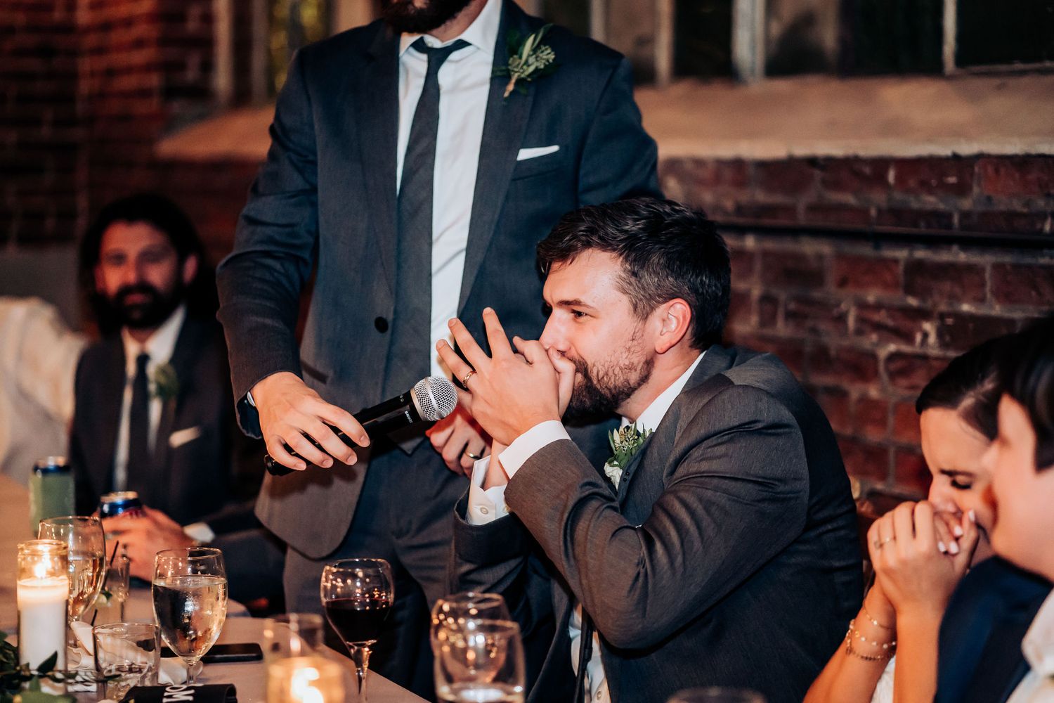 Wedding guests react emotionally during reception toasts at a rustic brick venue with candlelit tables.