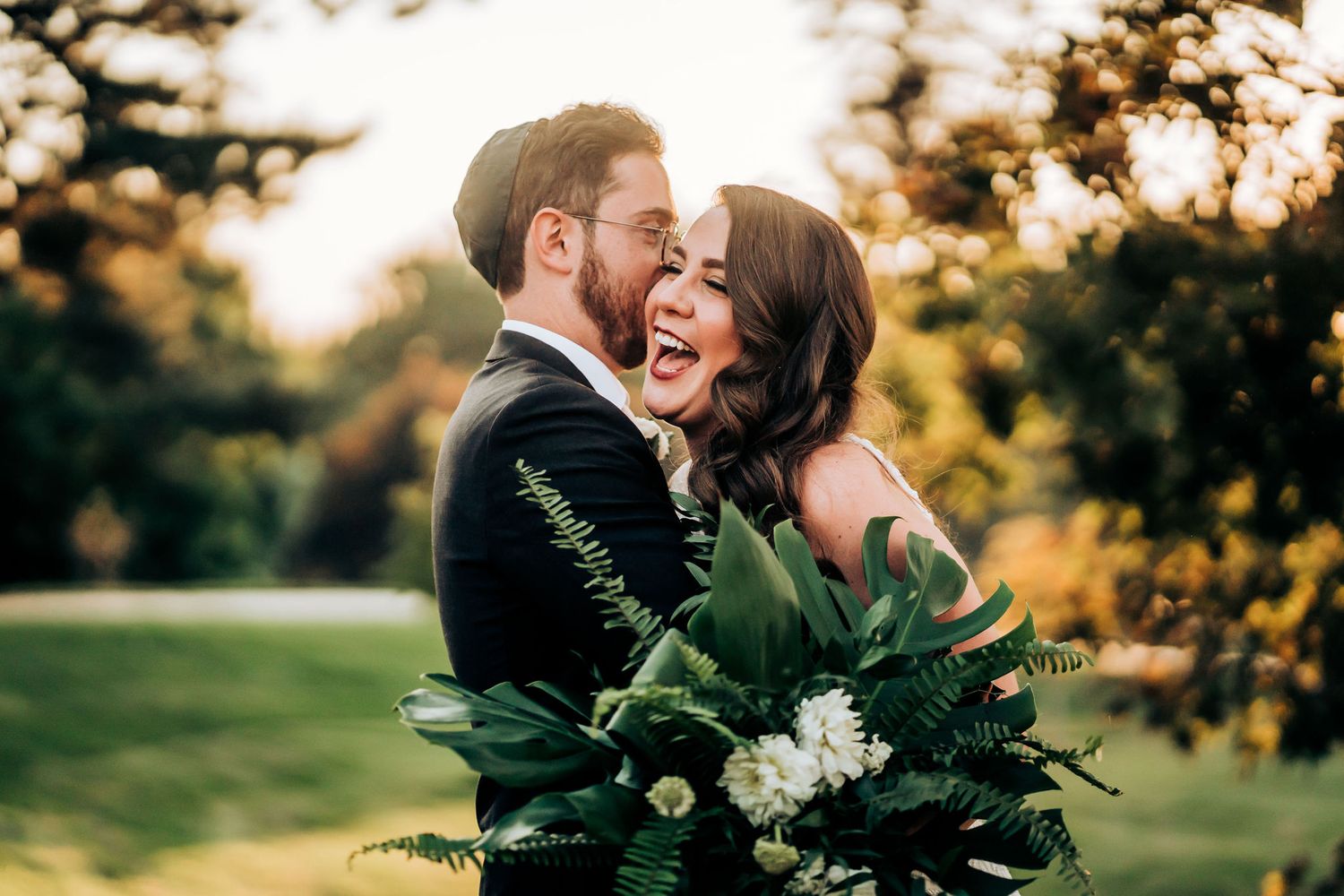 Joyful couple laughing during sunset portrait session with lush green fern bouquet.