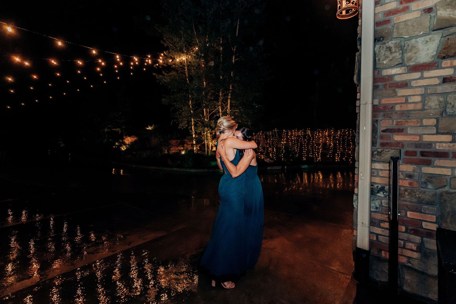 Person in blue dress stands beneath outdoor string lights at nighttime celebration.