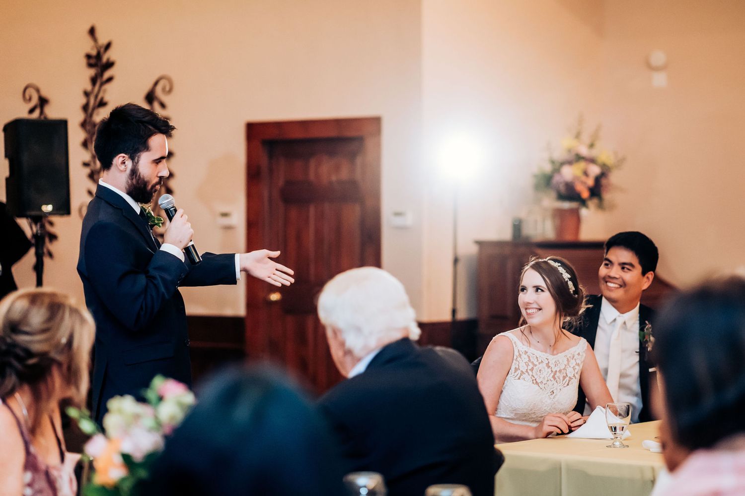 Speech being delivered during wedding reception in elegant indoor venue with decorative flowers.