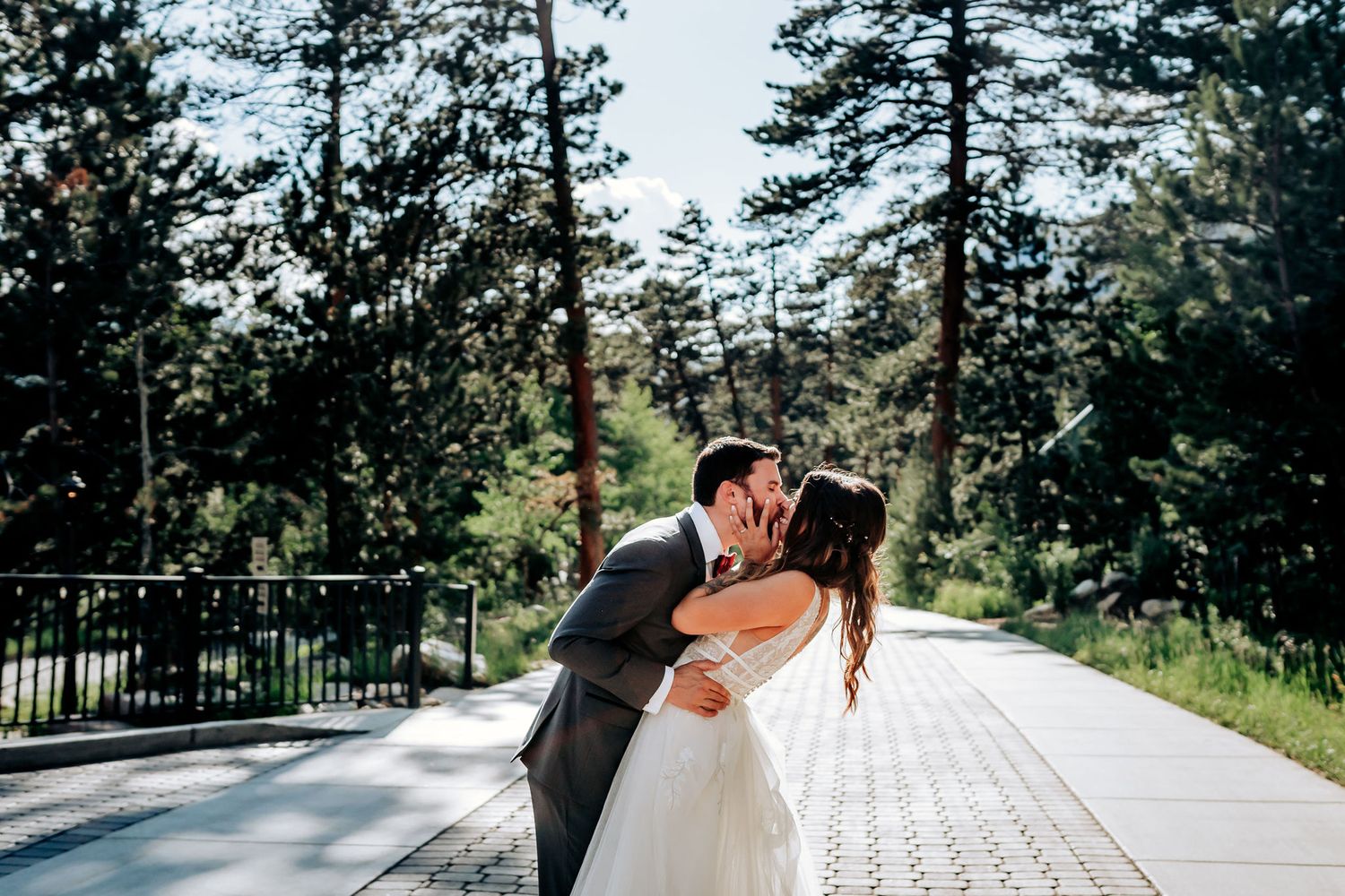 A romantic wedding couple embracing and kissing on an outdoor deck with pine trees in the background.
