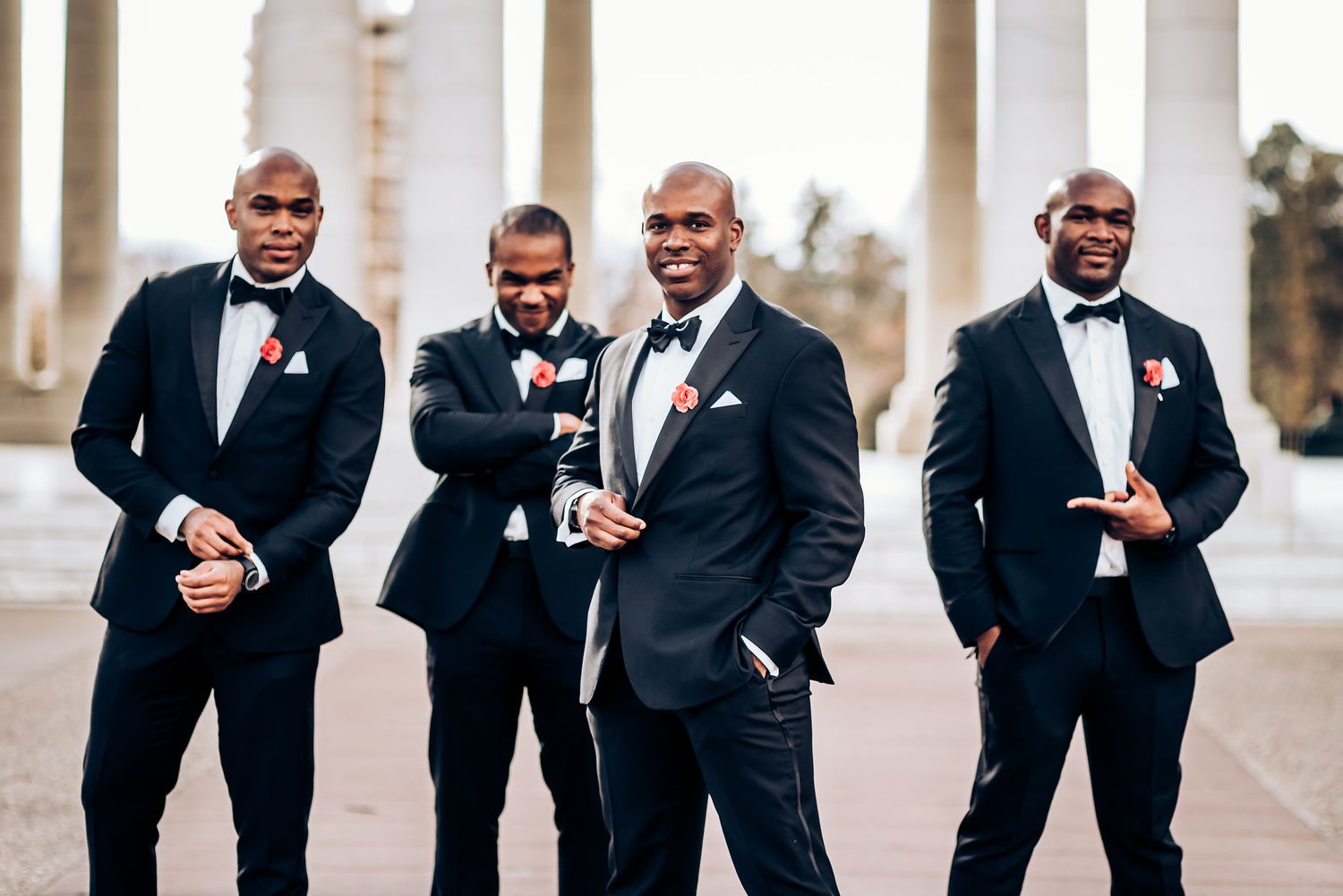 Groomsmen in black tuxedos share a lighthearted moment under classical columns.