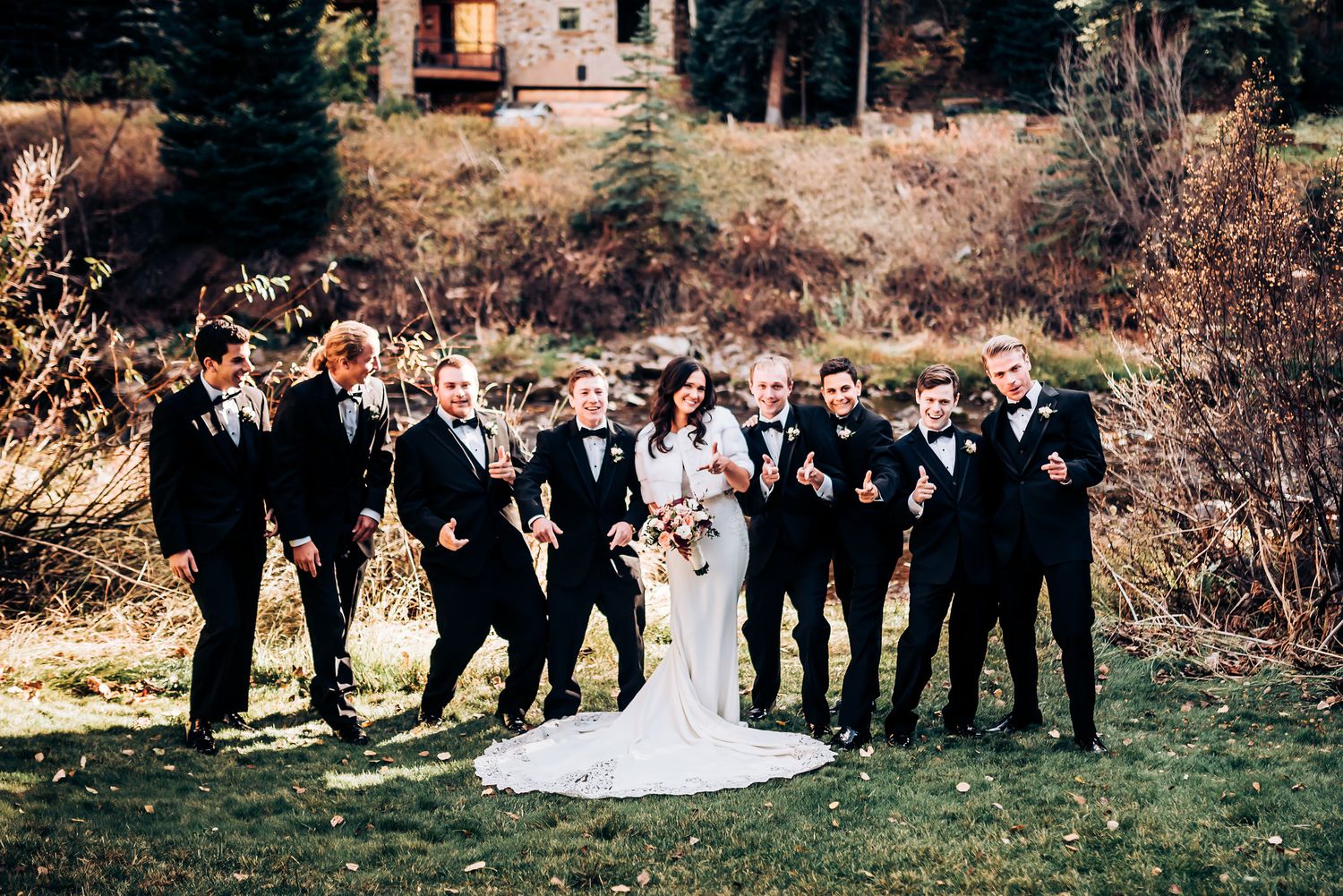 Groomsmen in black tuxedos pose with wedding couple on grassy lawn near stone building.