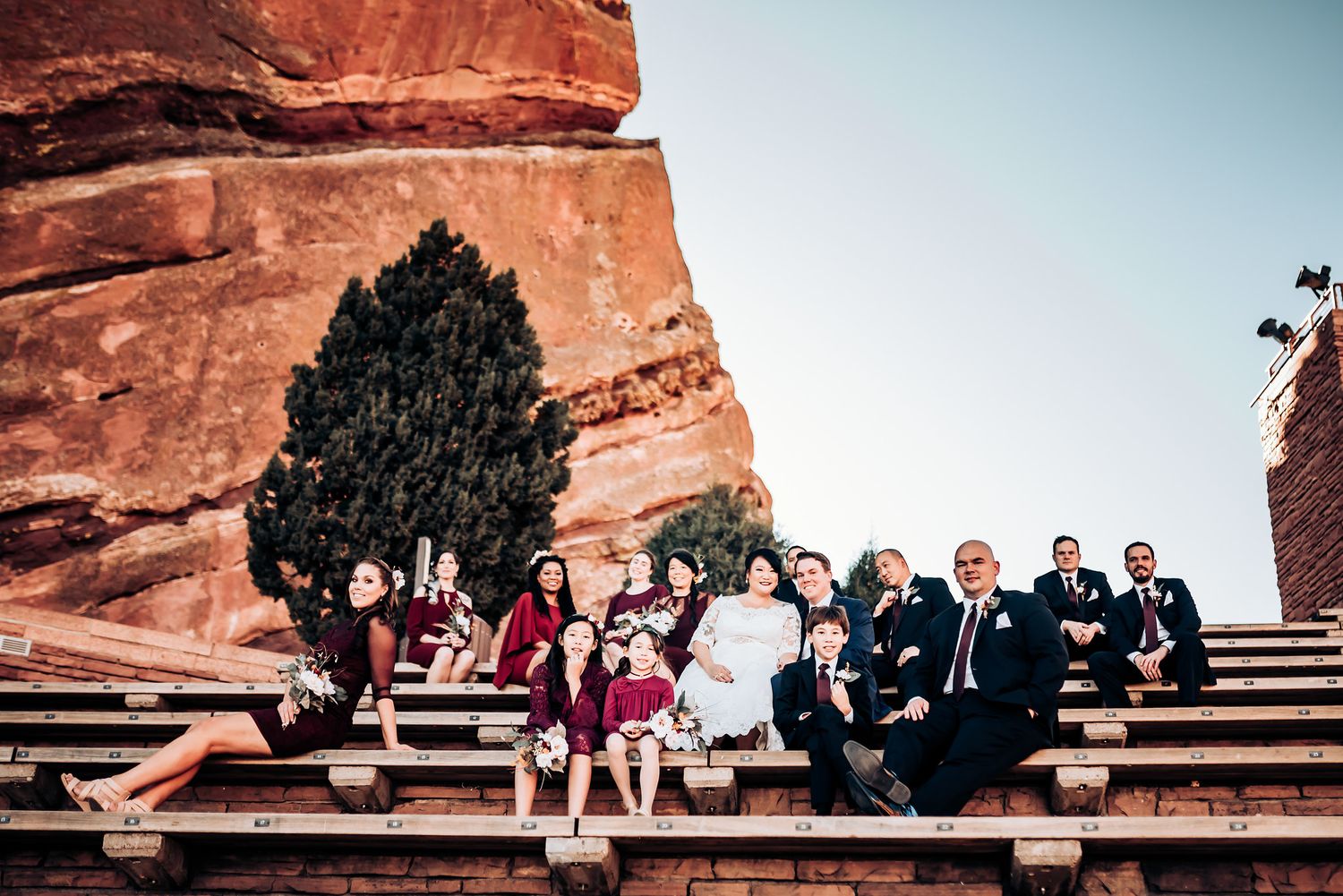 Wedding group seated on iconic Red Rocks amphitheater steps against blue sky.