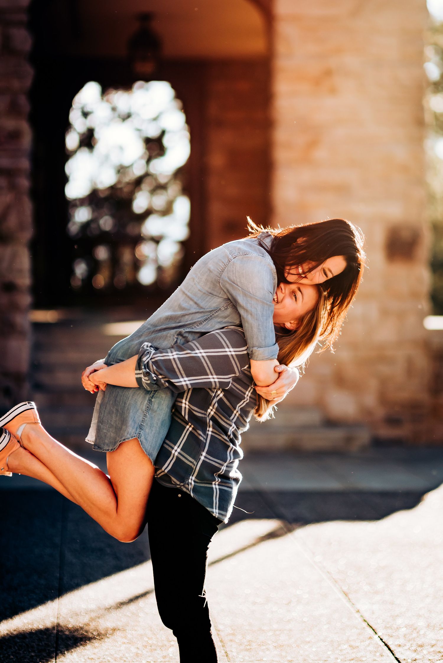 Playful moment captured as pair embraces in front of brick building with bokeh lighting.