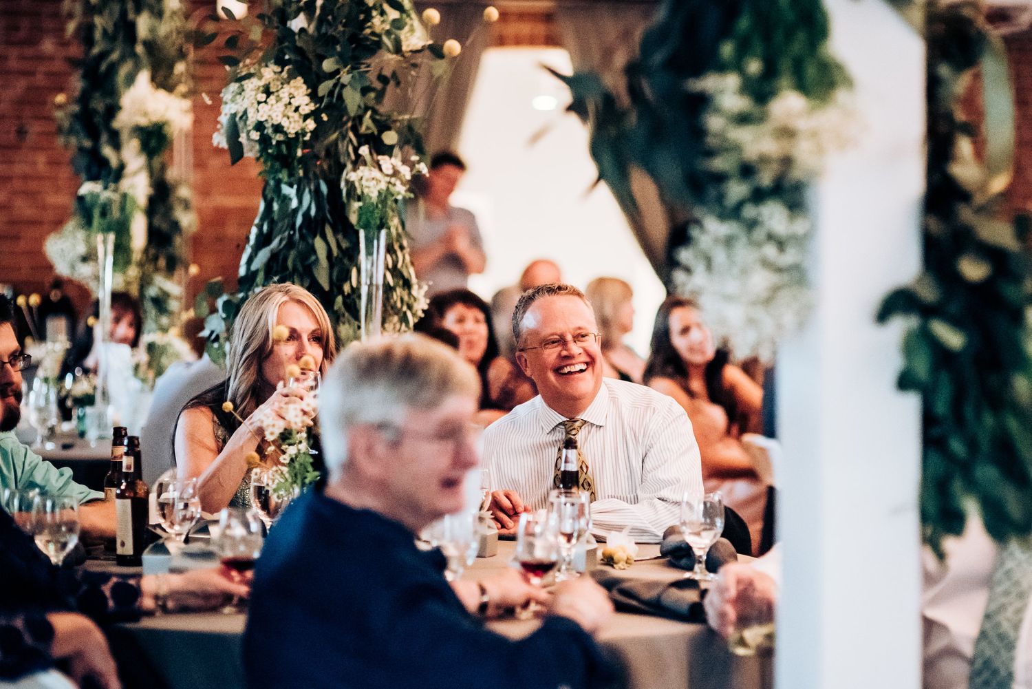 Wedding reception guests laugh and smile around a decorated table with floral arrangements.