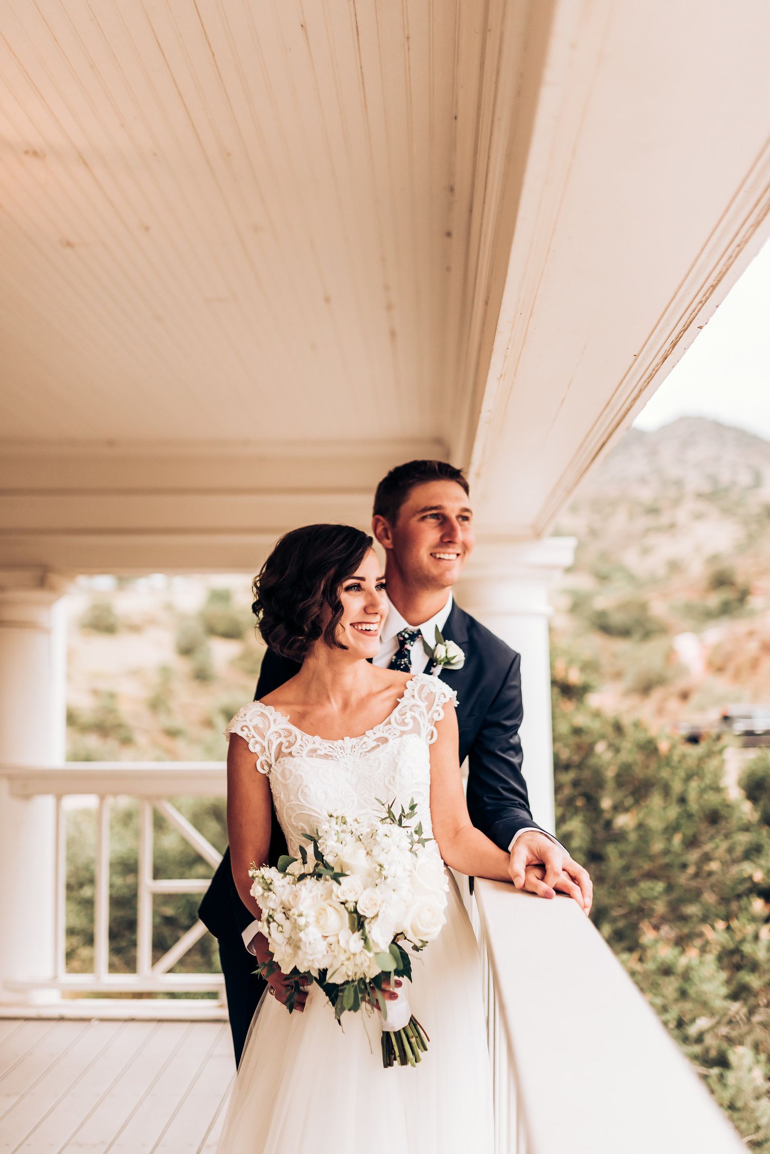 A couple in wedding attire stands on an elegant white porch overlooking mountain scenery.