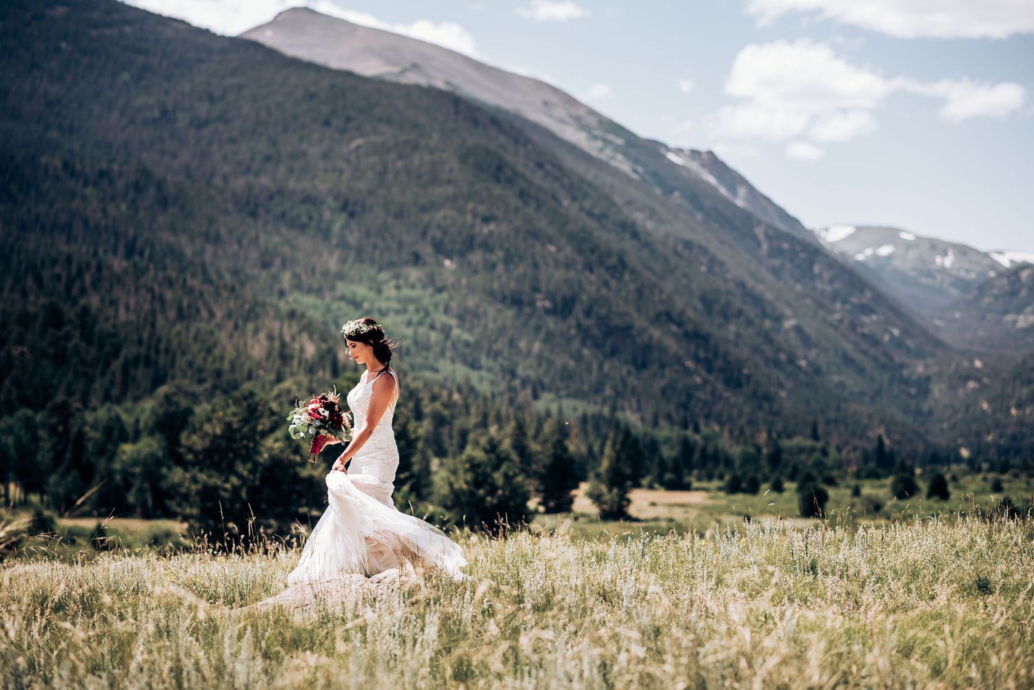 A flowing white wedding dress catches the mountain breeze in a sunny alpine meadow.