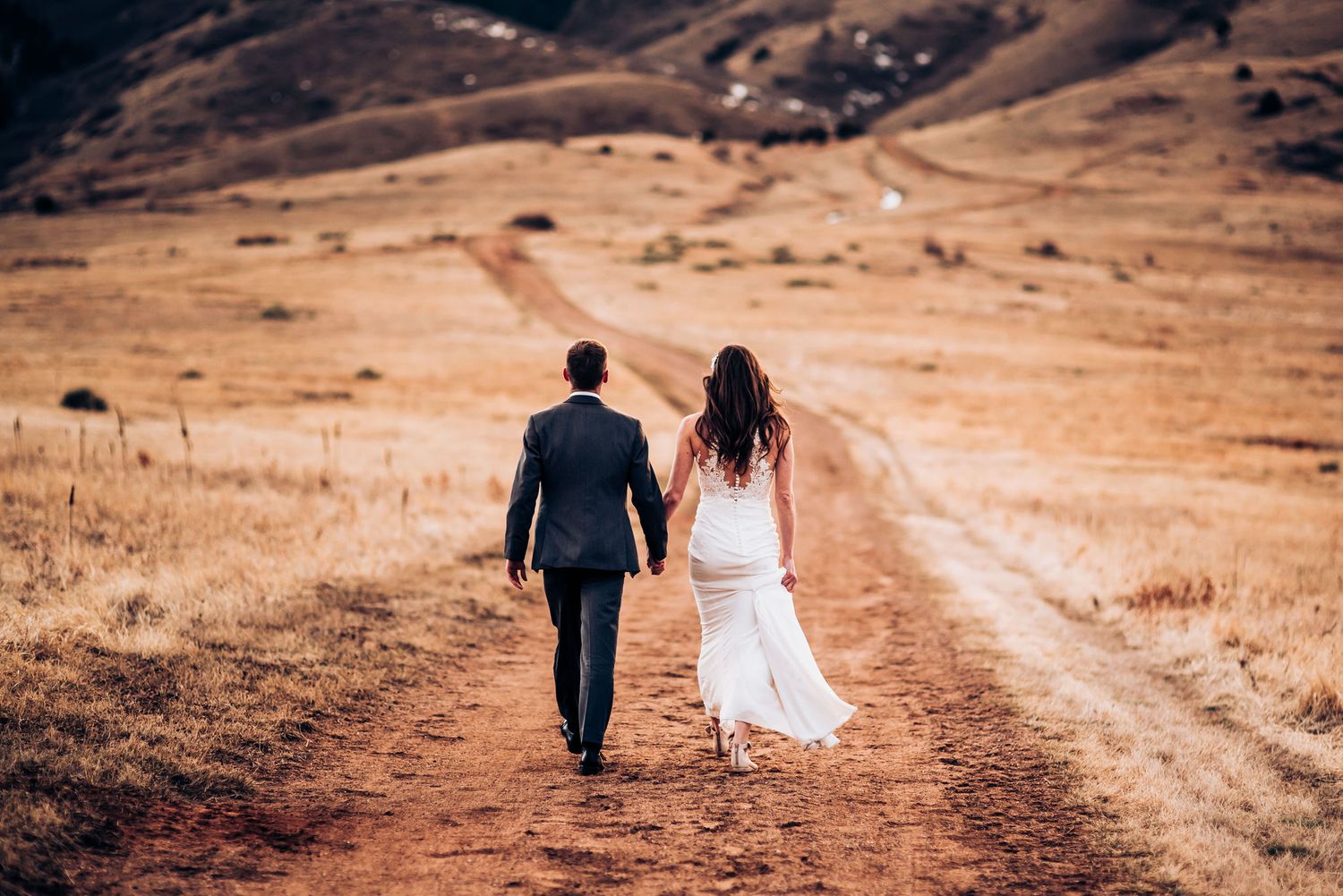 Wedding couple walking hand in hand down rustic mountain path at sunset.