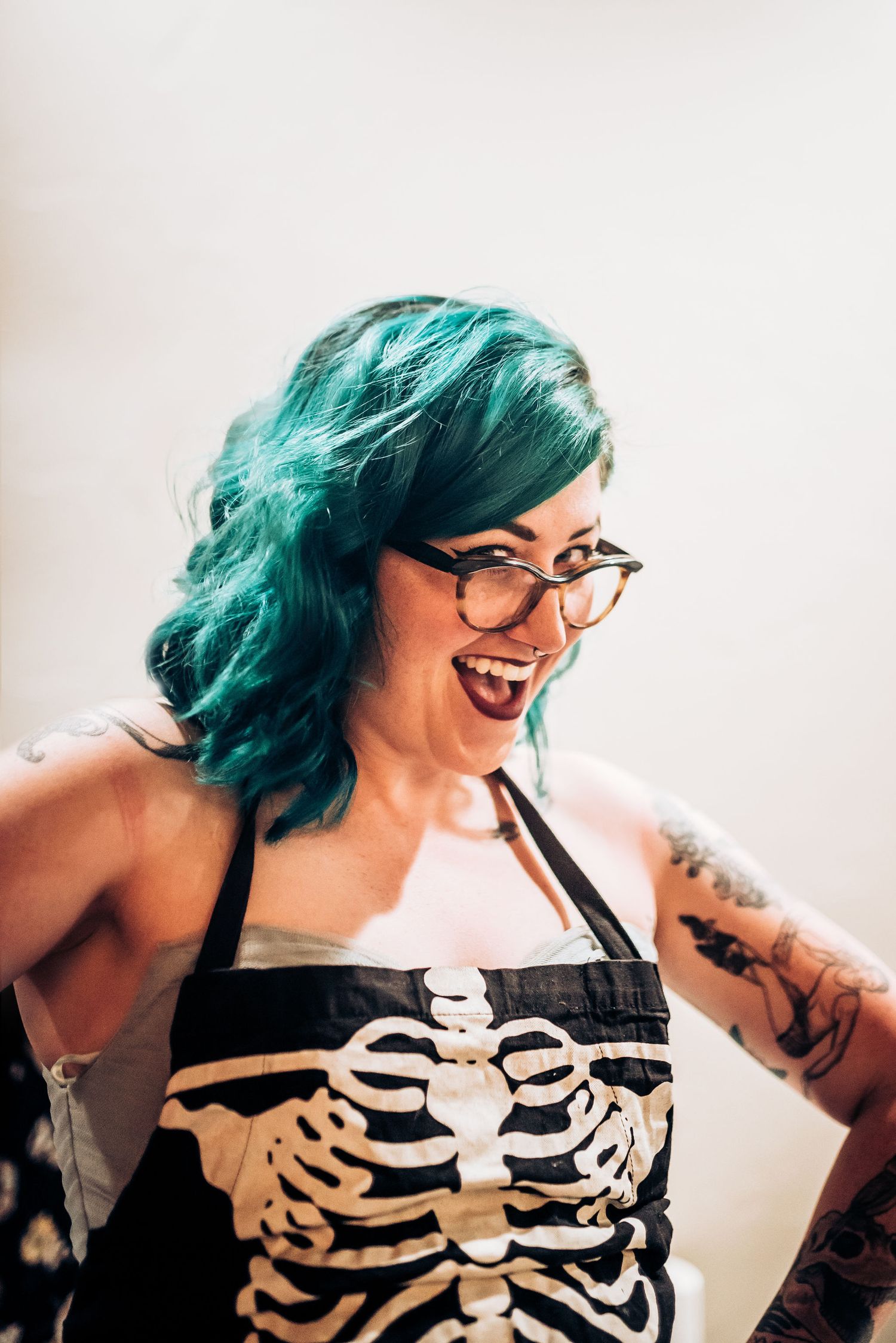 Woman with teal hair wearing skeleton print top and glasses smiling brightly.