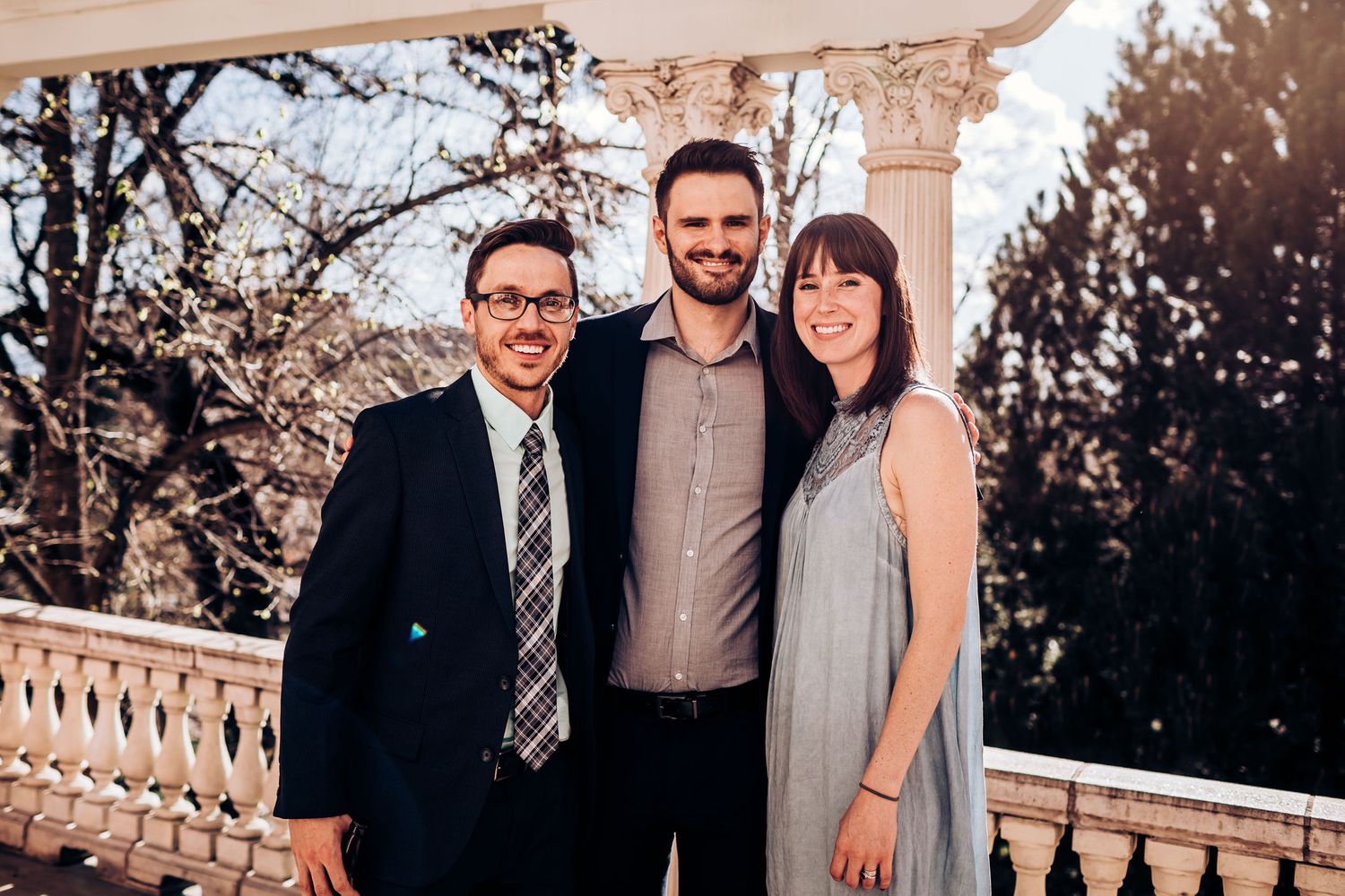 Three guests pose together on an elegant balcony with ornate architecture and trees in background.