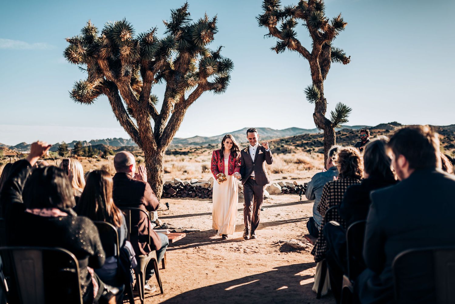 Wedding guests watch ceremony under majestic Joshua Trees in a desert setting.