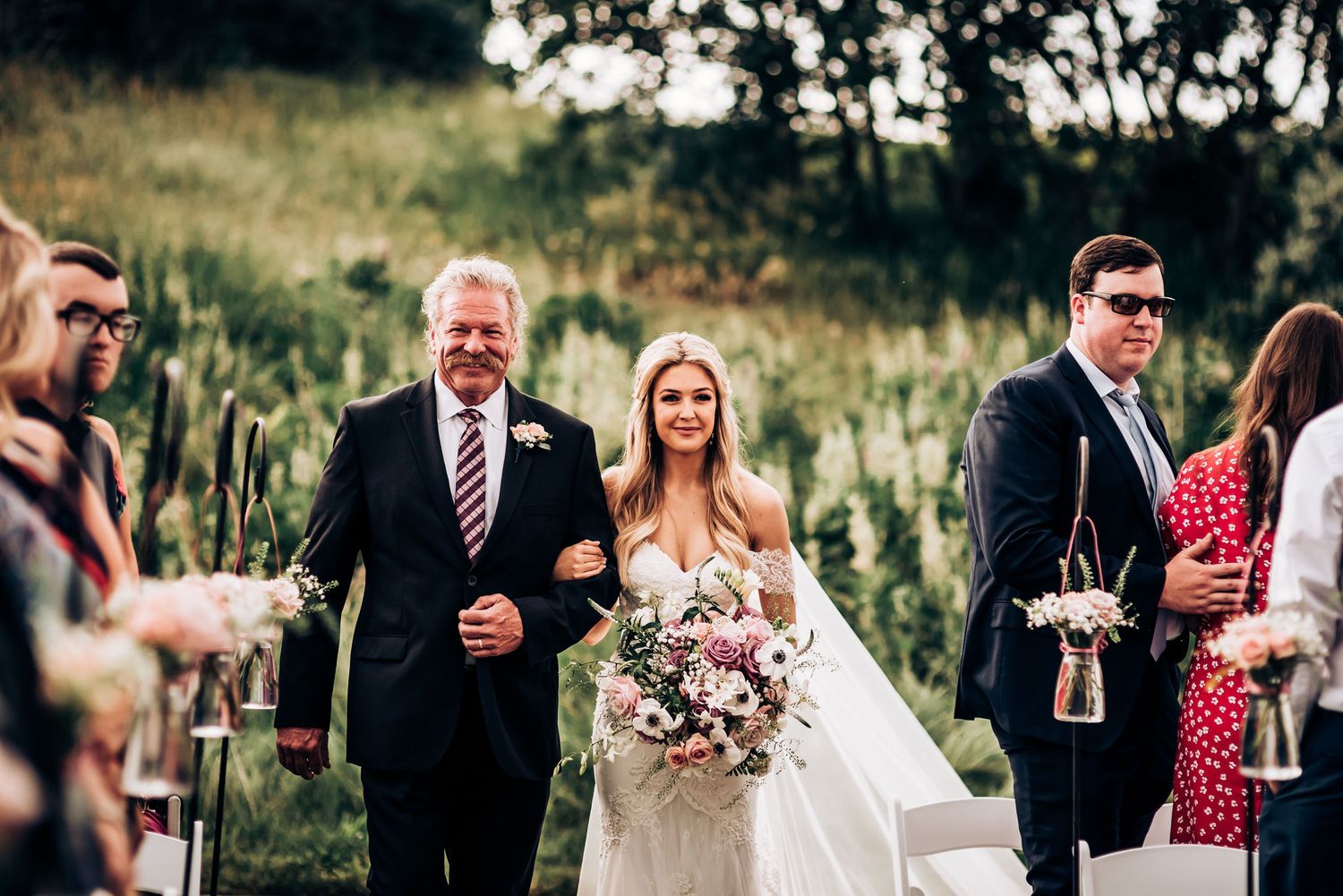 Wedding procession walks down outdoor aisle with bouquet of pink flowers.