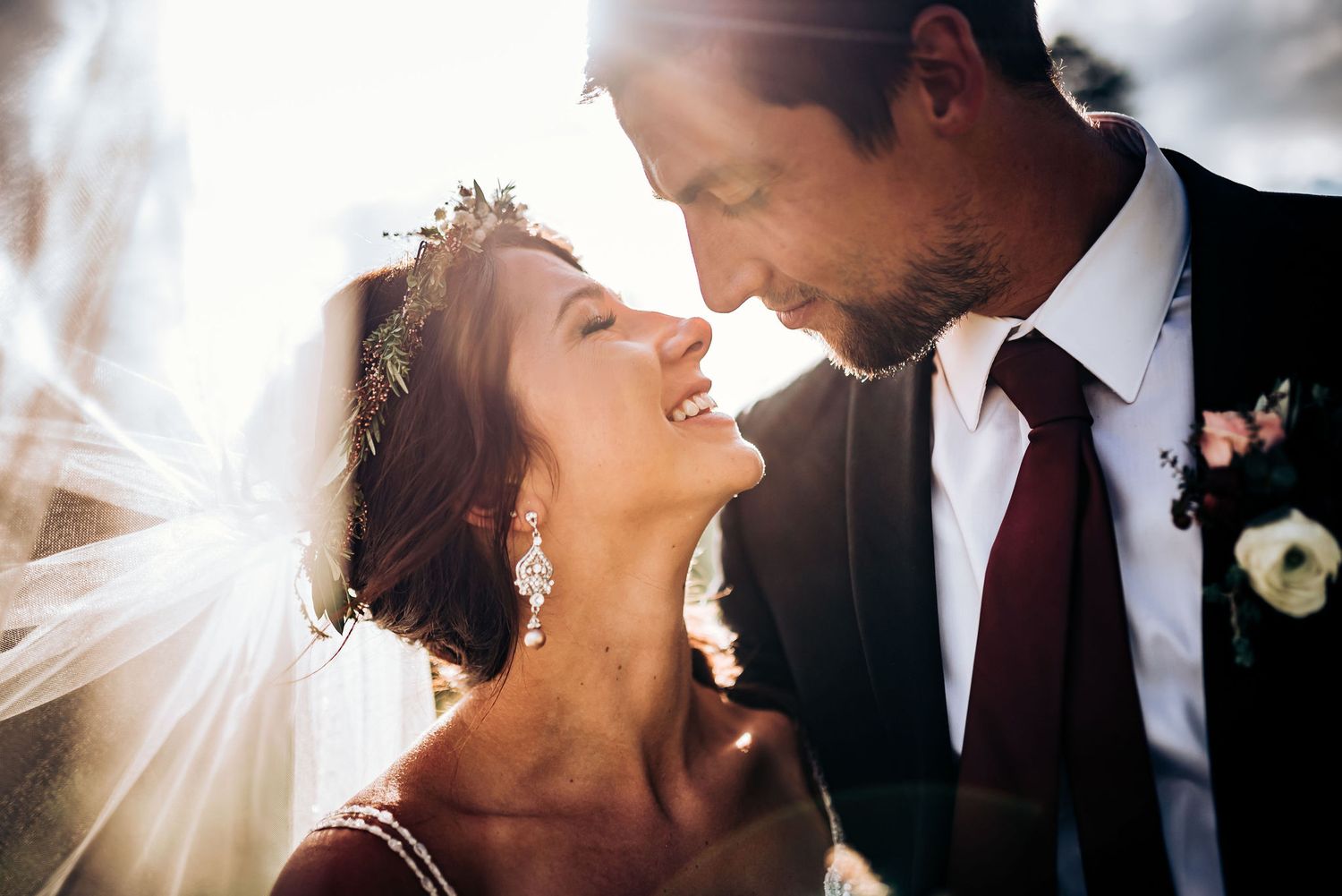 Romantic wedding portrait of couple sharing an intimate moment with sunlit veil and burgundy accents.