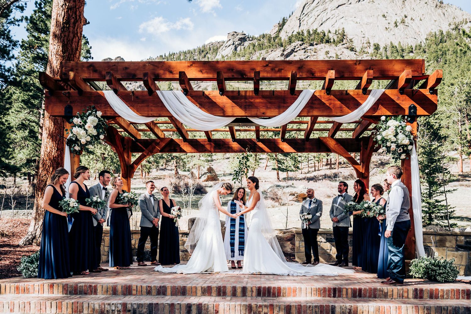 Wedding ceremony gathering under wooden pergola with mountain peak in background.
