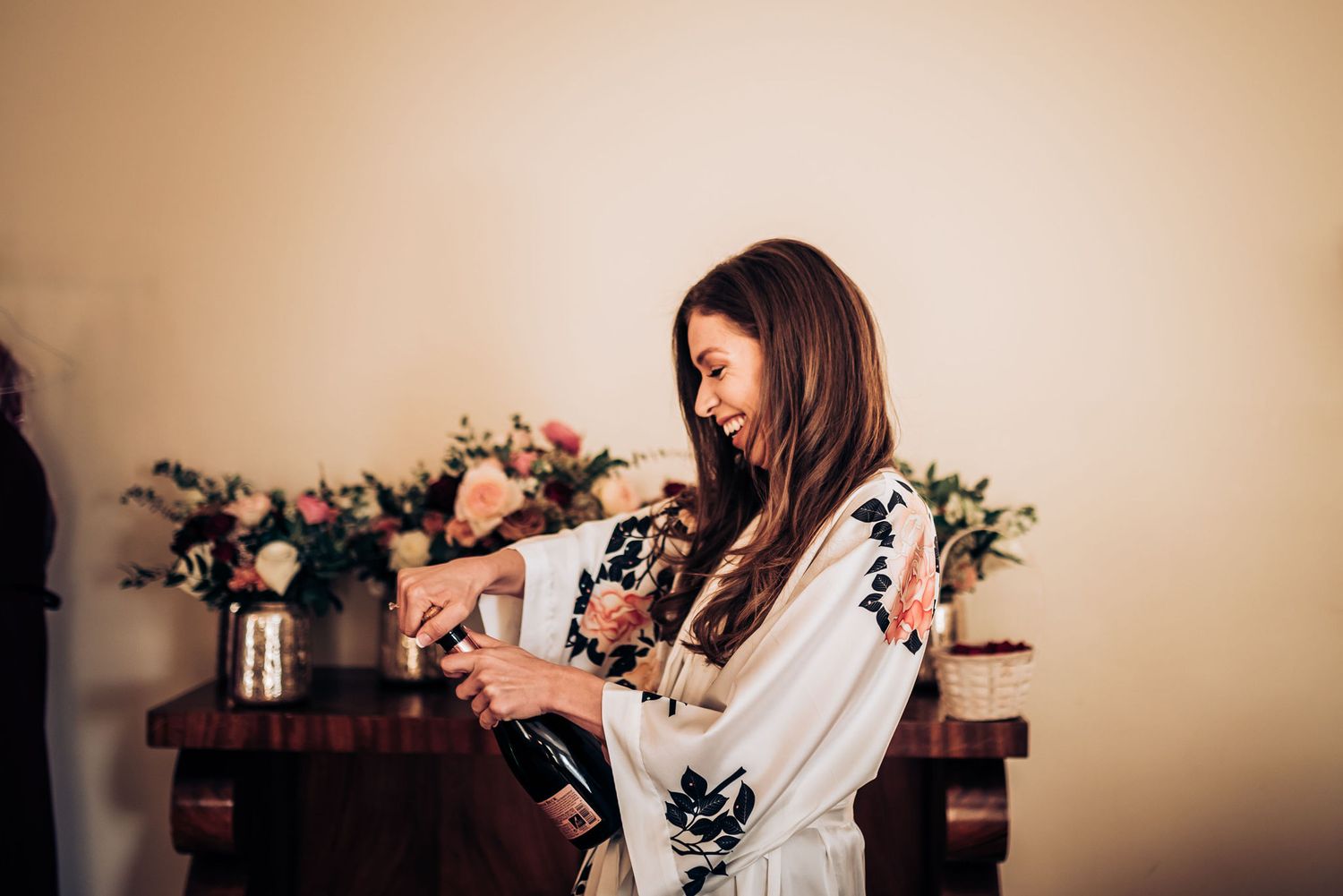 Someone in floral robe pours champagne during wedding morning preparations.