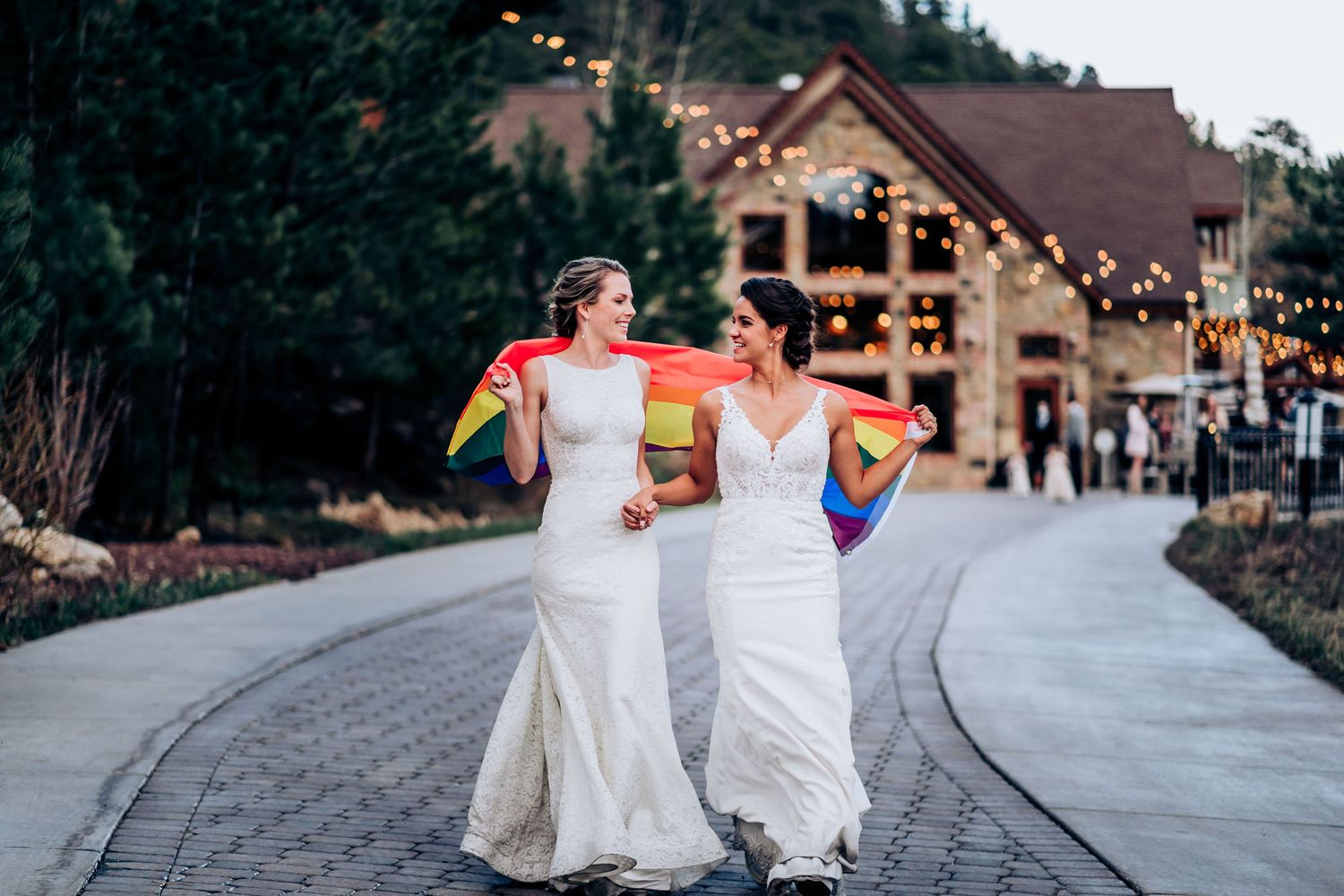 Two people in wedding dresses walk together holding rainbow flags outside a venue with string lights.