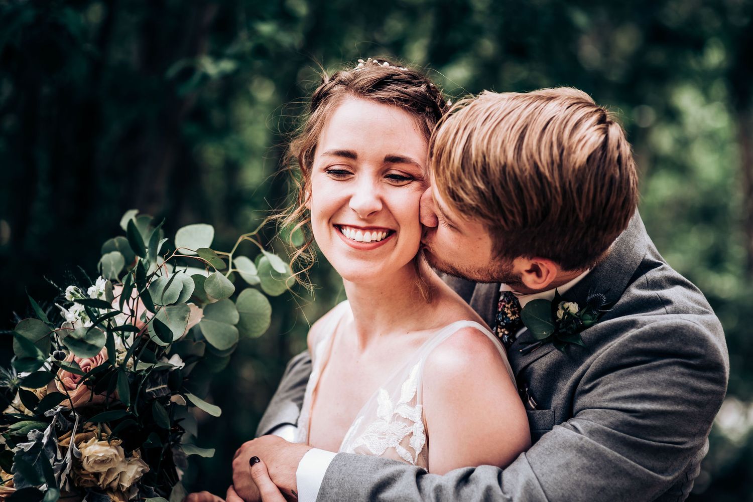 Intimate moment between newlyweds with greenery and flowers in an outdoor forest setting.