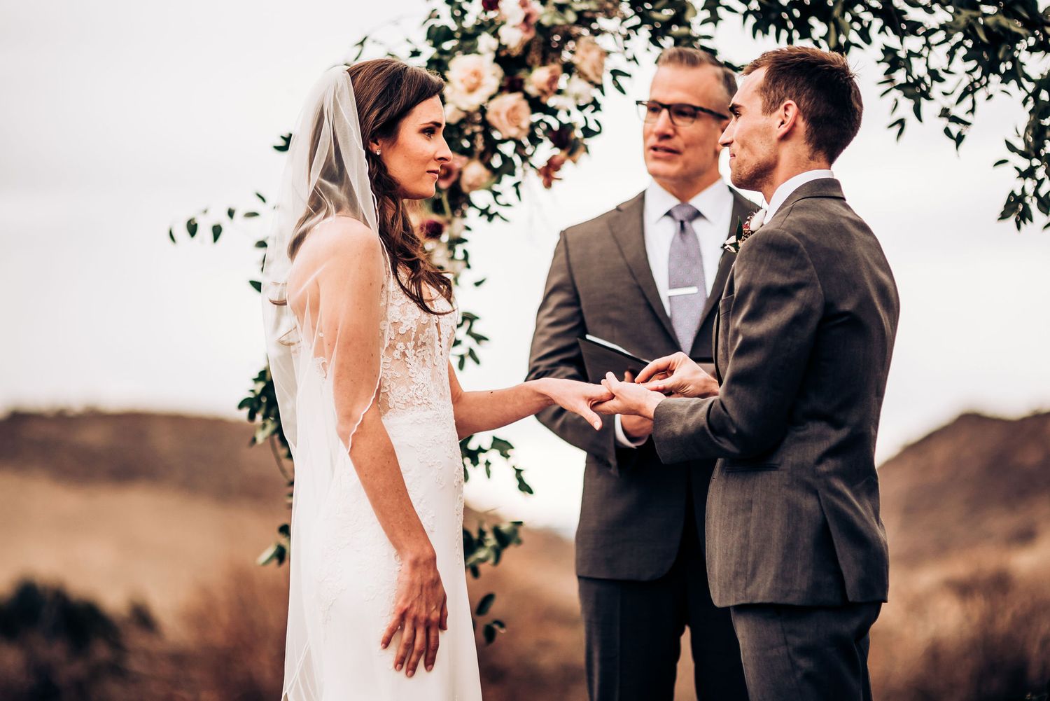 Exchange of rings during an outdoor wedding ceremony beneath a floral arch.
