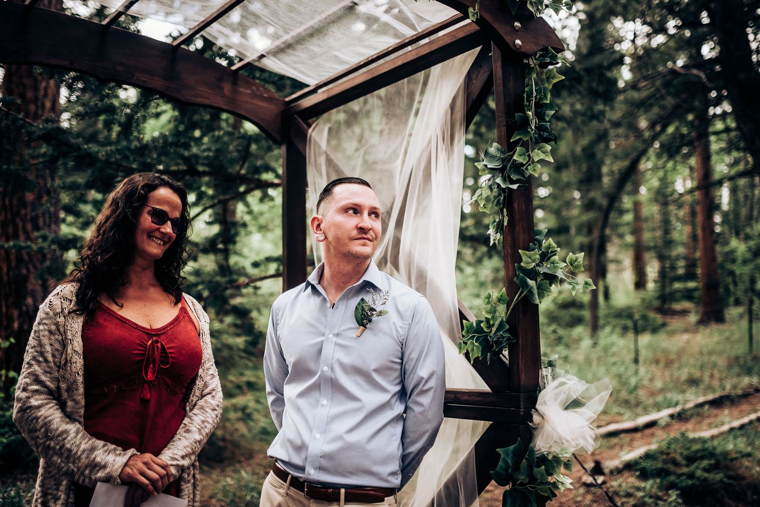 A couple stands together under a wooden ceremony arch in a forest setting.