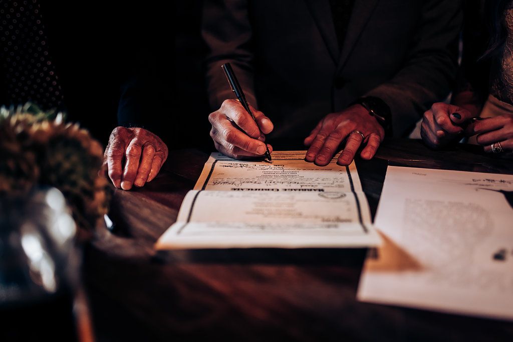 Close-up shot of hands signing official wedding documents with a floral arrangement visible.
