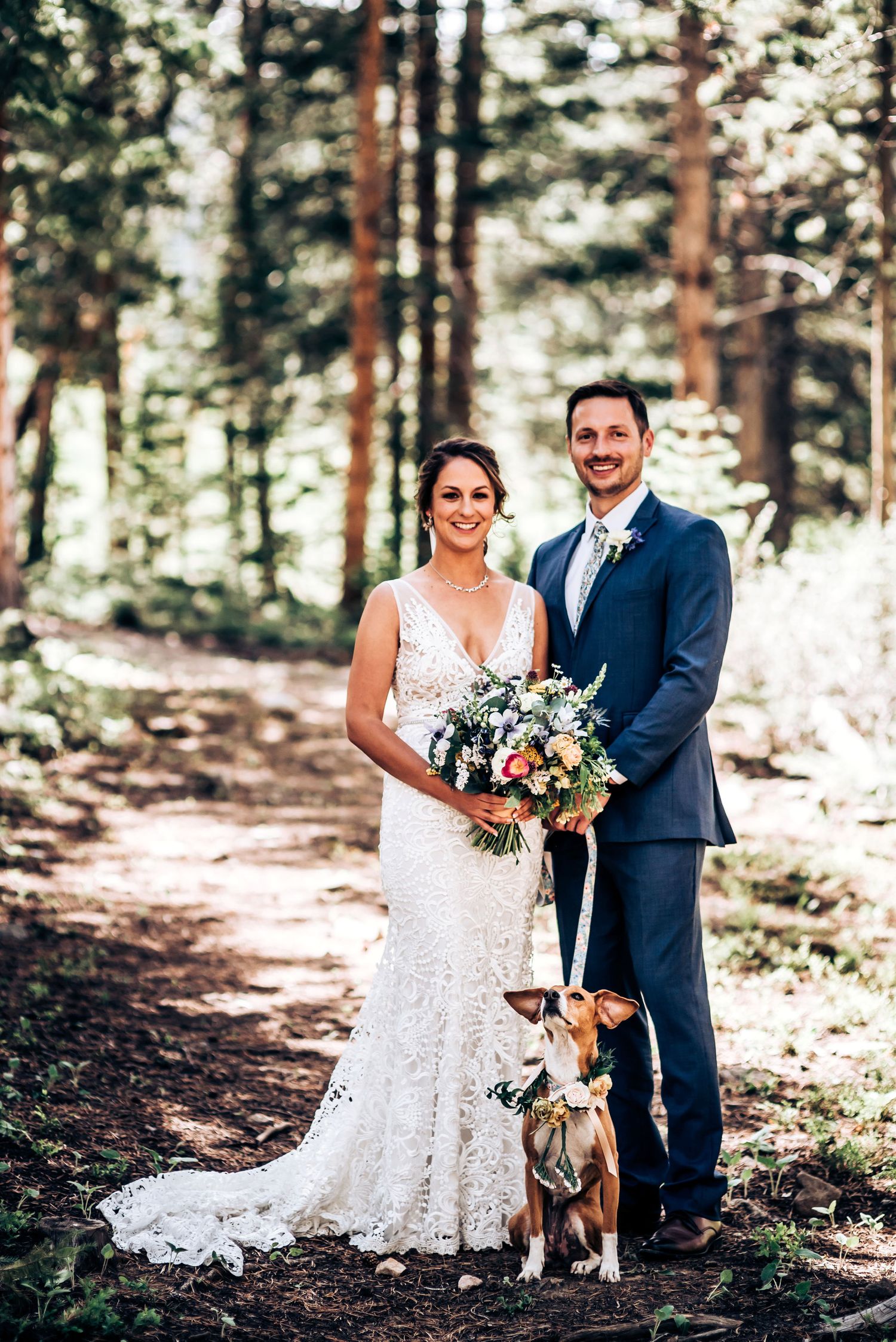 A wedding couple poses with their dog on a forest trail wearing formal attire.